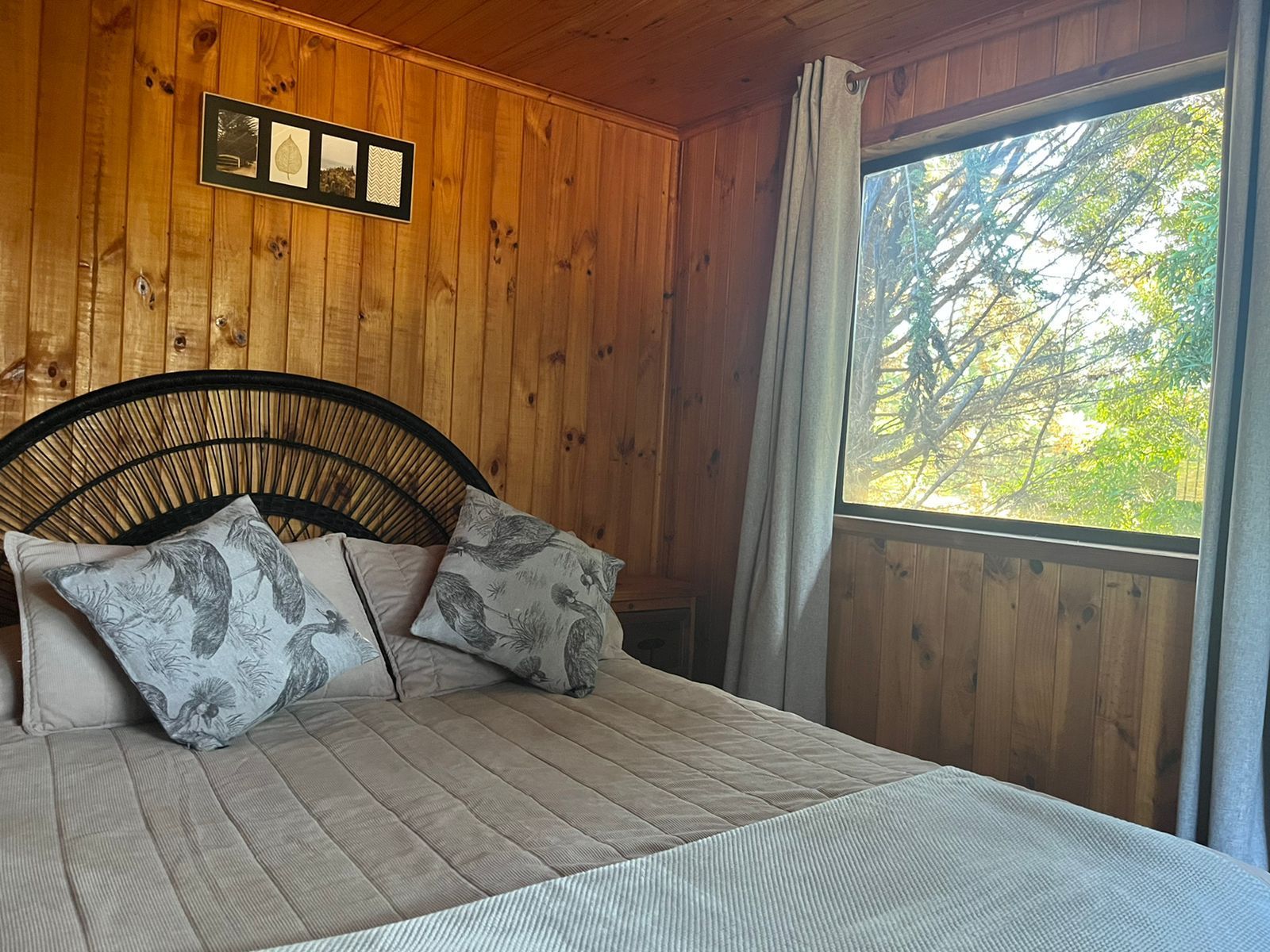 Dormitorio con cama, revestimiento de madera, cabecero de mimbre y ventana con cortinas ligeras.