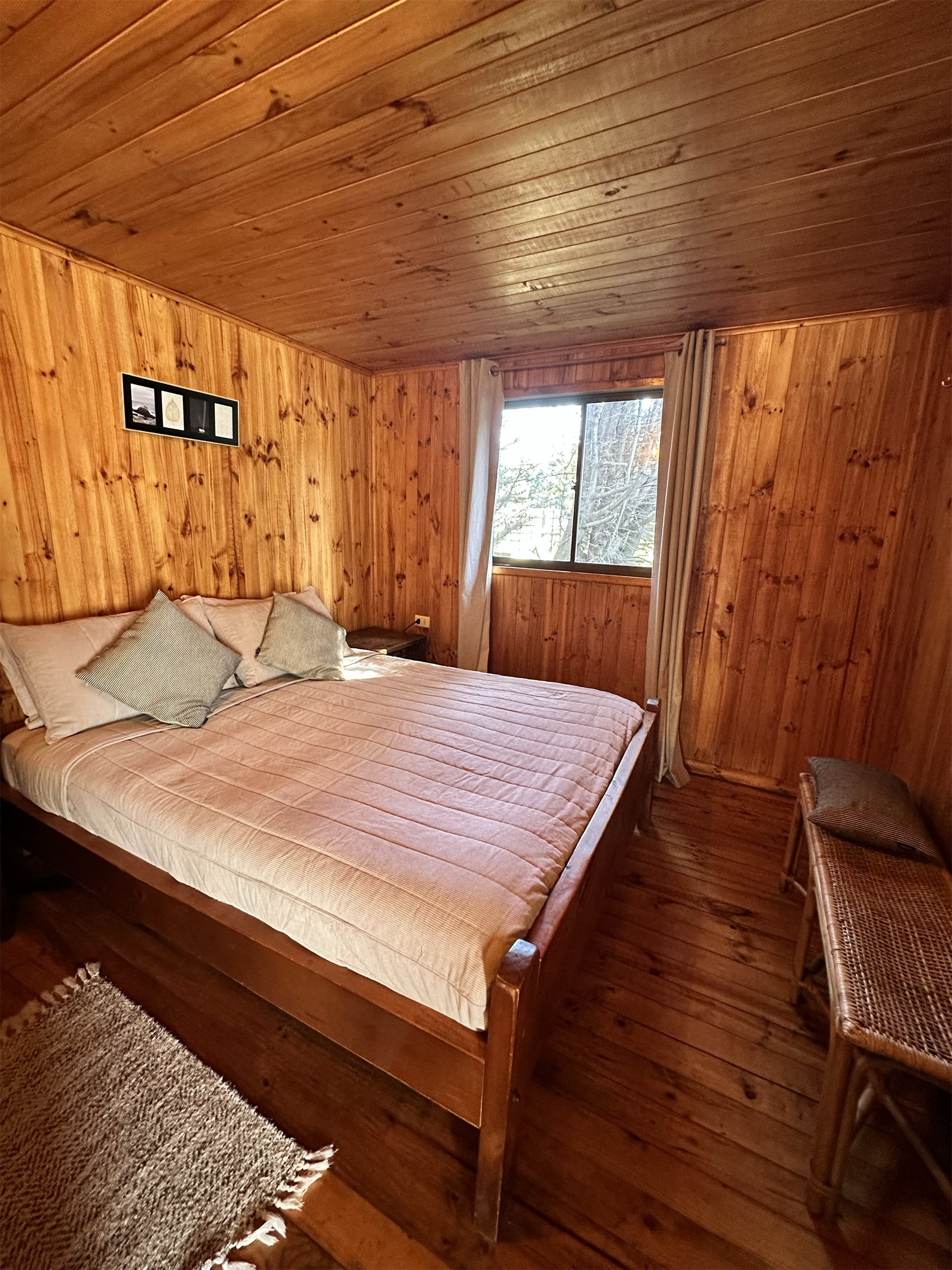 Dormitorio con paredes y techo de madera, cama con almohadas, pequeña ventana, banco y alfombra.