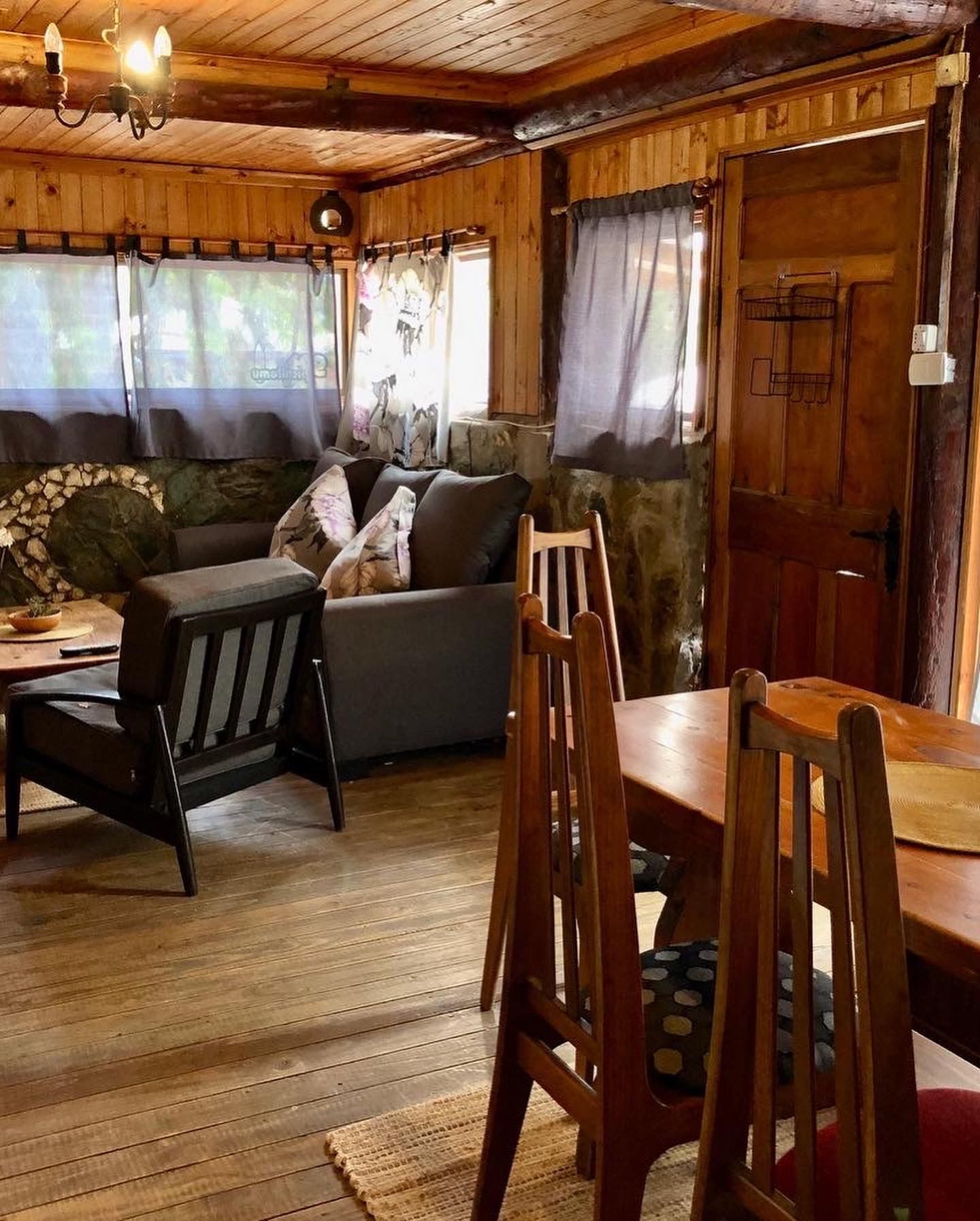 Acogedor interior de cabaña revestido de madera: sala de estar con sofá, sillas, mesa de comedor y ventanas con cortinas.
