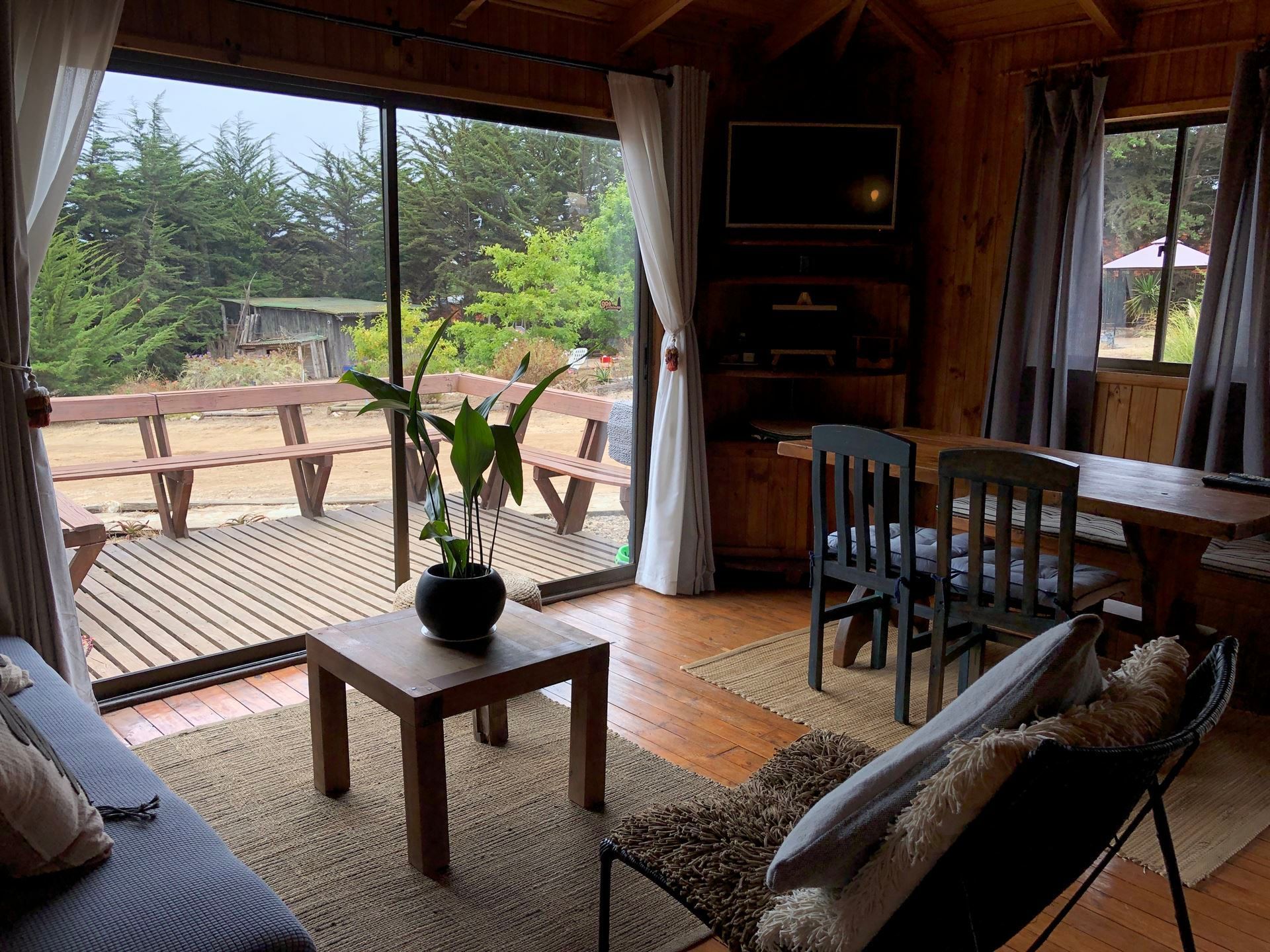 Acogedor interior de cabaña con amplios ventanales con vistas a la terraza. Sala de estar con sofá, mesa y comedor.