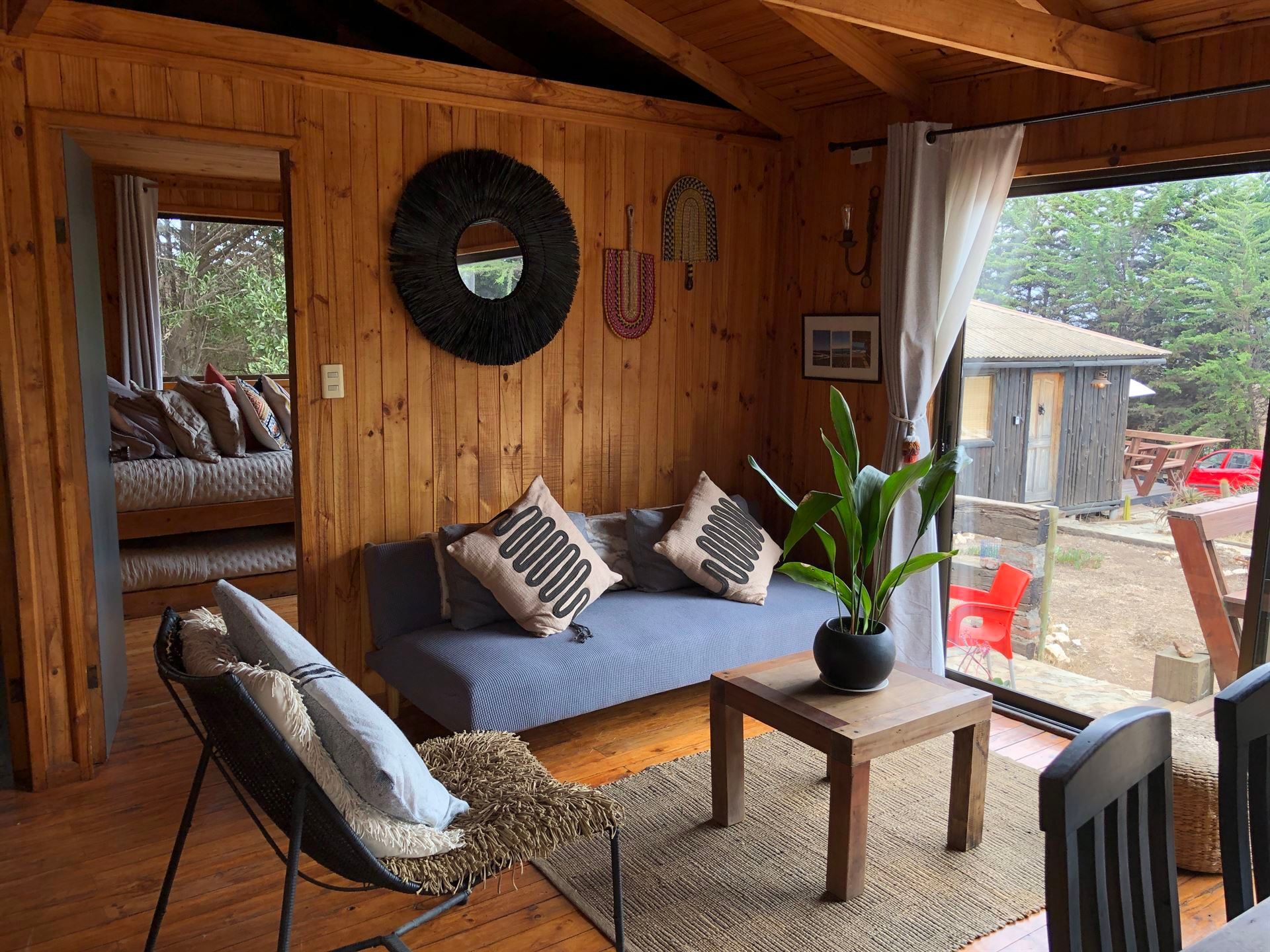 Acogedor interior de cabaña con paneles de madera, sofá, sillas y una gran ventana con vista a un patio.