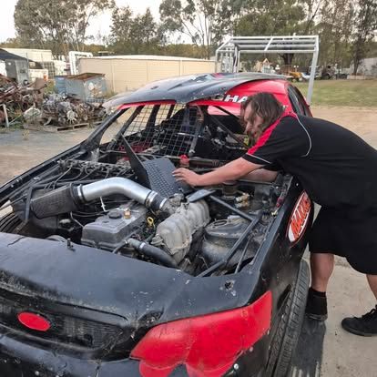Man working on the engine of a heavily modified black race car with red accents in a junkyard.— Genesis Automotive Services in Lavington, NSW