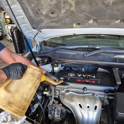 Mechanic pouring oil into a Toyota engine bay. Open hood, black gloves, tan oil container.— Genesis Automotive Services in Lavington, NSW