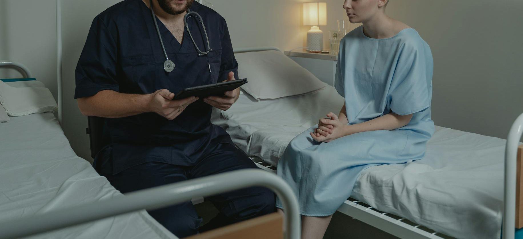A doctor in scrubs consults with a patient in a hospital room. The patient wears a hospital gown.