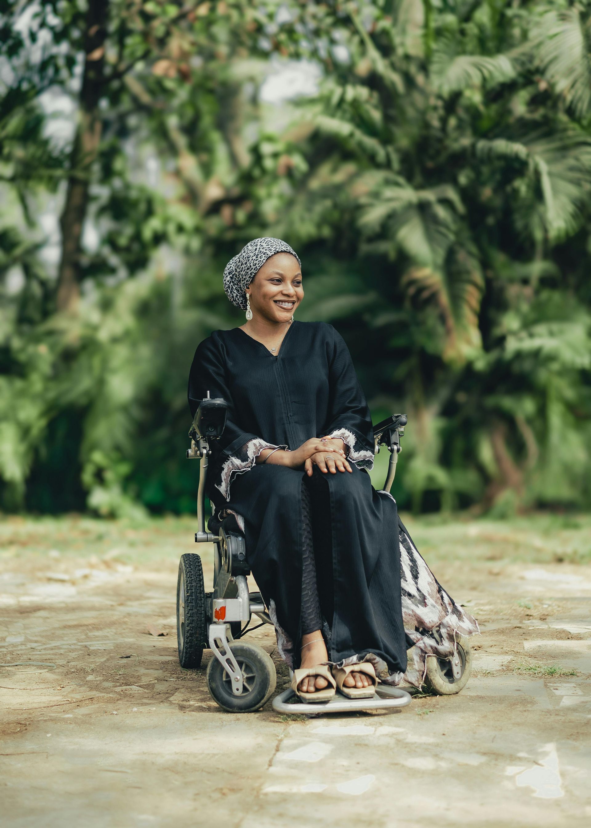 Woman in a black dress and headscarf sits in a wheelchair outdoors, smiling in a natural setting.
