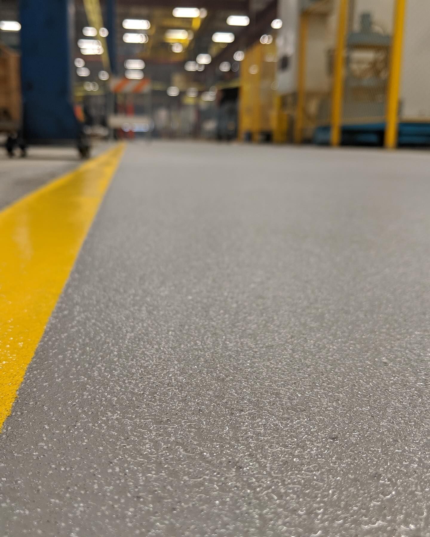 Close-up of a gray industrial floor with a yellow painted border. Blurred factory setting in the background.