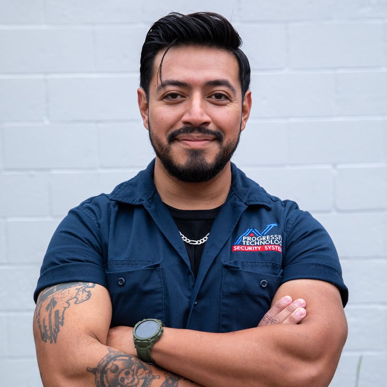 Man with crossed arms, wearing a dark blue mechanic shirt, smiling, against a white brick wall.