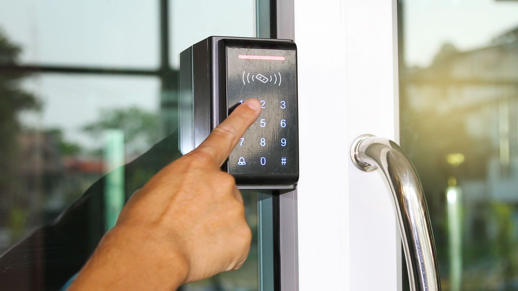 Person's hand entering code on a keypad door lock on a glass door, in a building.