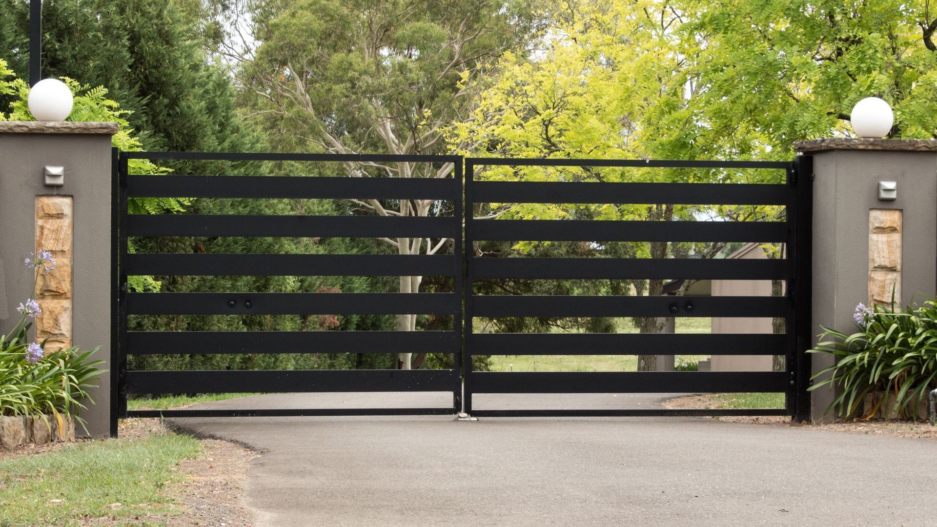 Black horizontal slat driveway gates, gray pillars with lights, asphalt driveway.