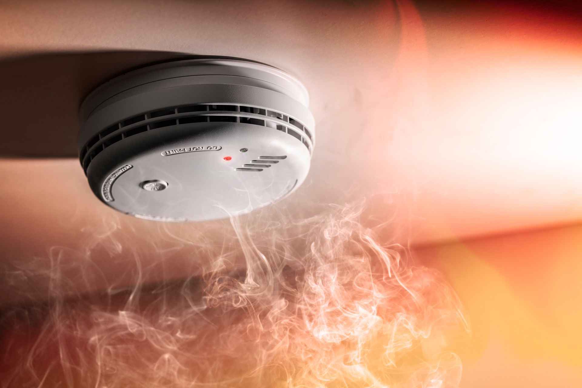 Smoke detector on a ceiling, with red light illuminated, smoke billowing below.