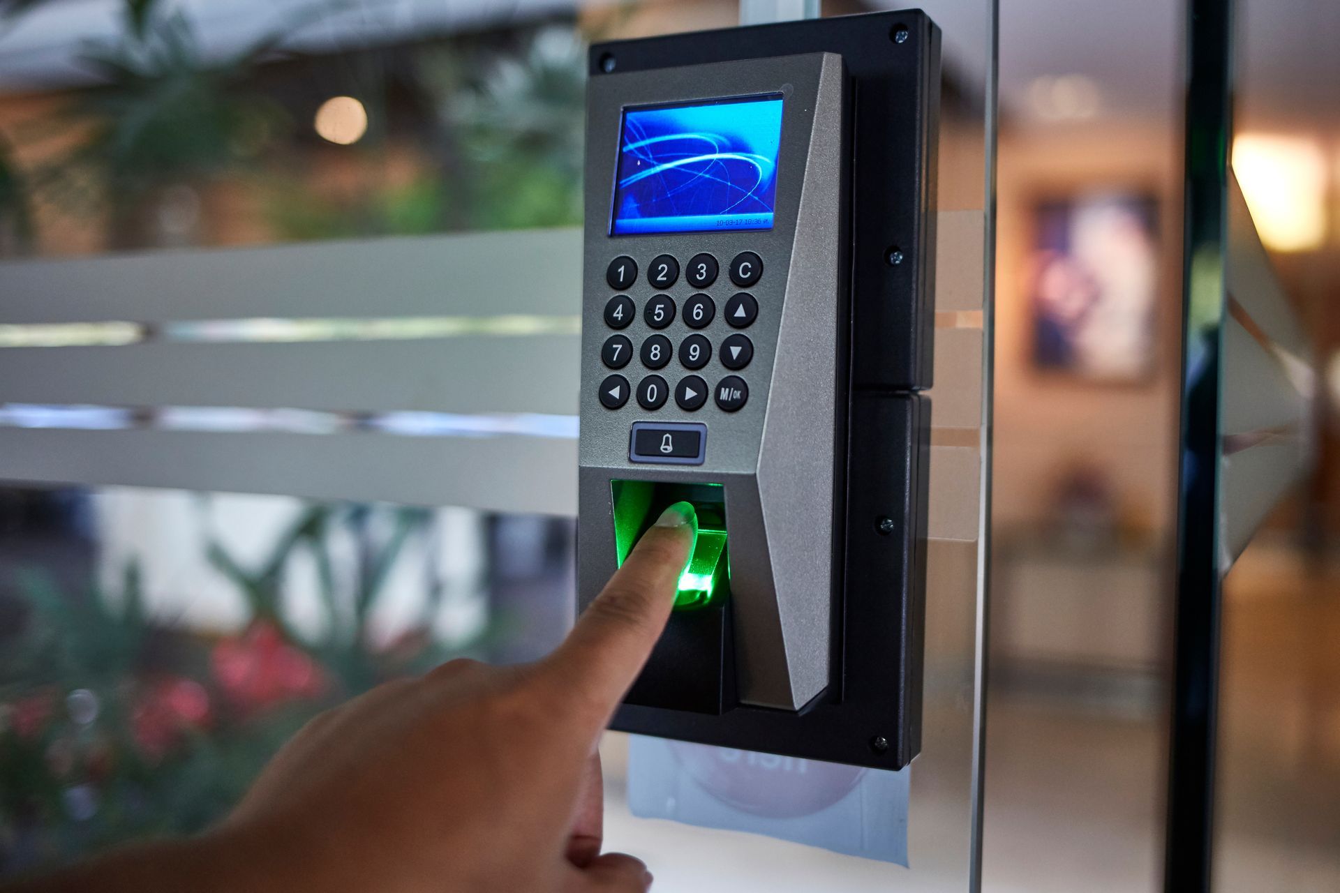Person swipes a card at a black door access system on a glass door; green foliage visible in the background.