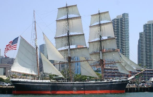 Tall ship with billowing white sails and an American flag, docked in a harbor with city buildings.
