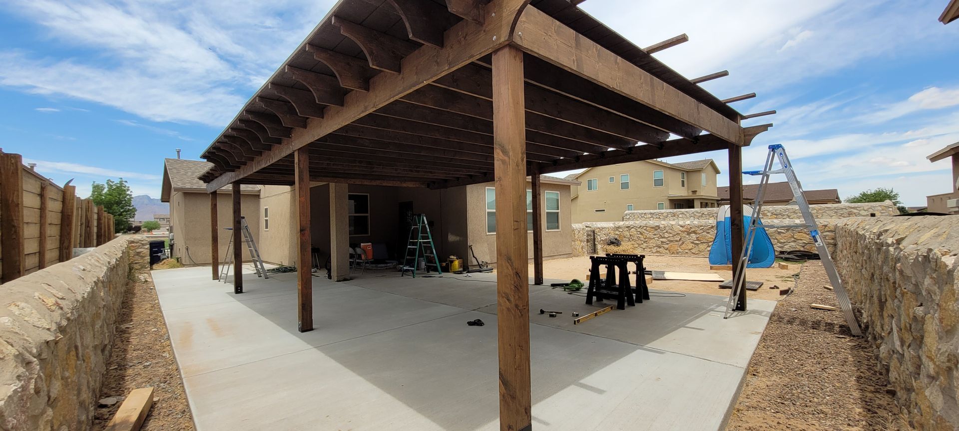 A wooden pergola covers a concrete patio with a view of a neighborhood under a cloudy sky.