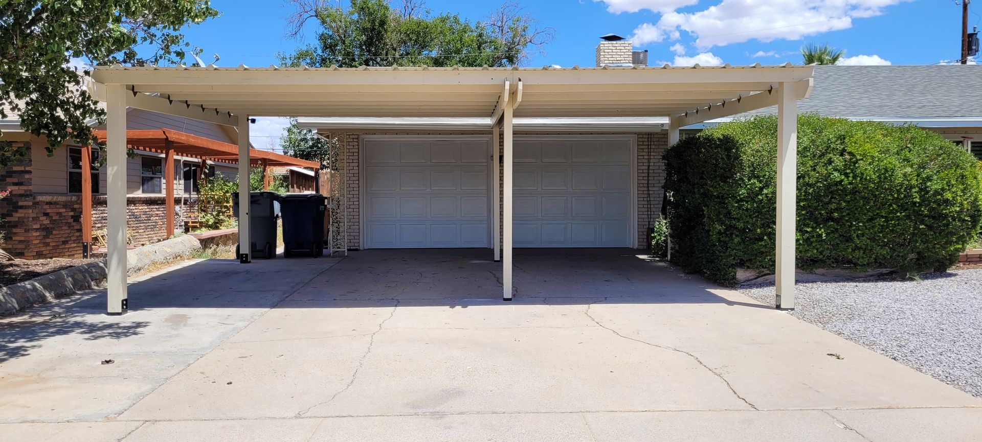 A carport with a two-car garage under a blue sky. Green bushes and trees are present.