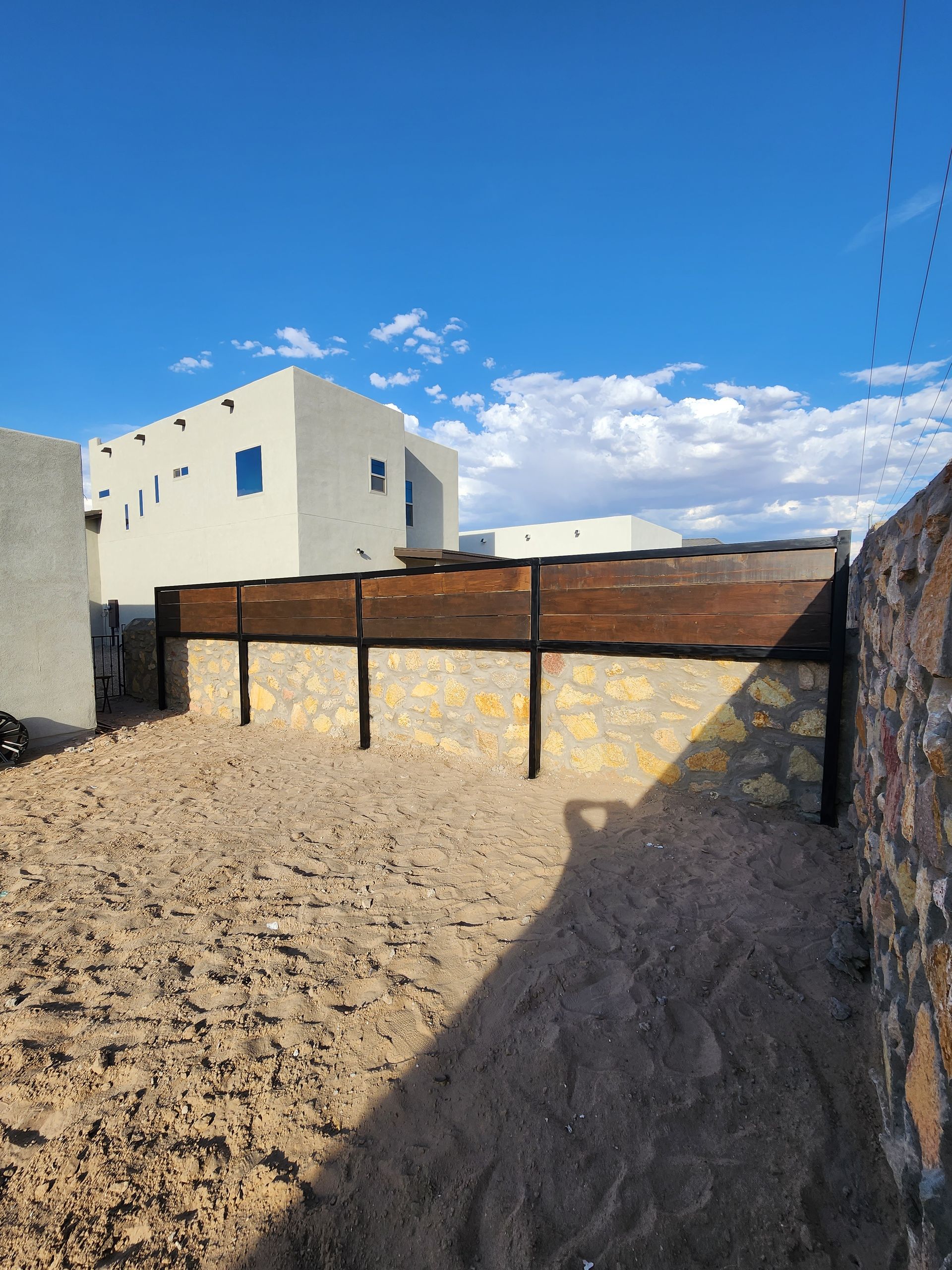 Sandy yard with a stone and wood fence under a blue sky.