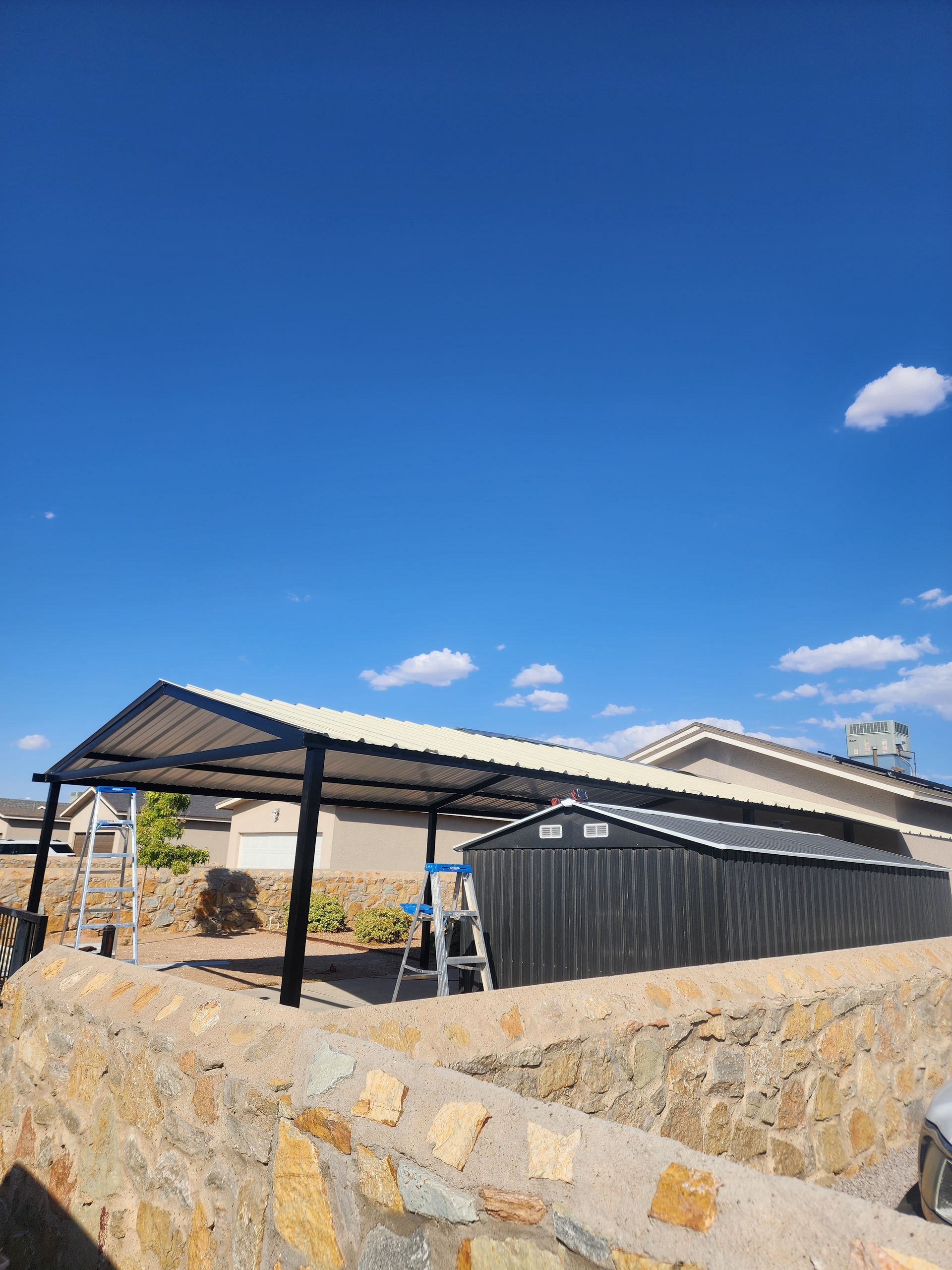 A metal carport with a shed in a sunny setting against a bright blue sky.