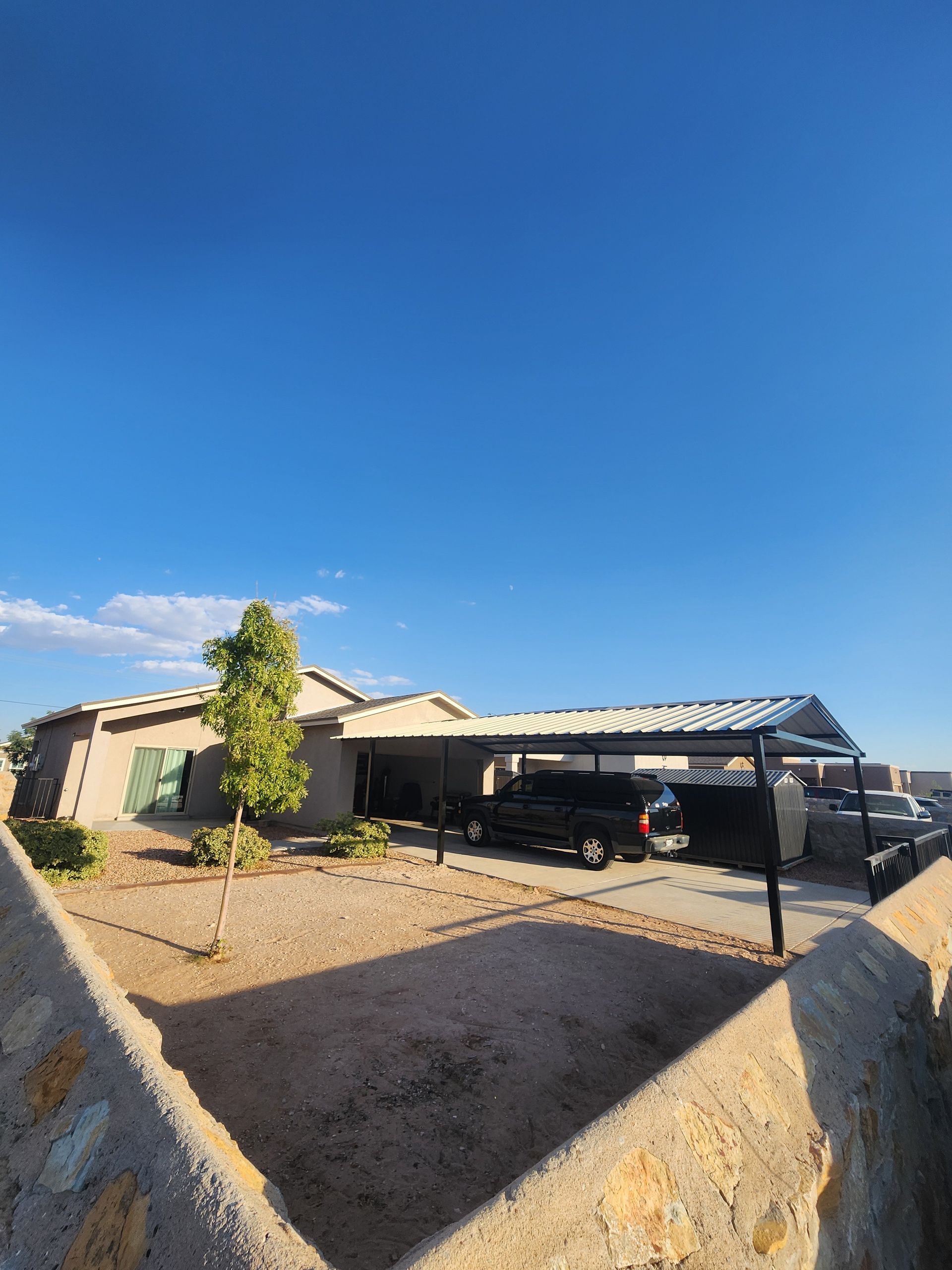 A single-story house with a carport, parked cars, a blue sky, and a low stone wall.