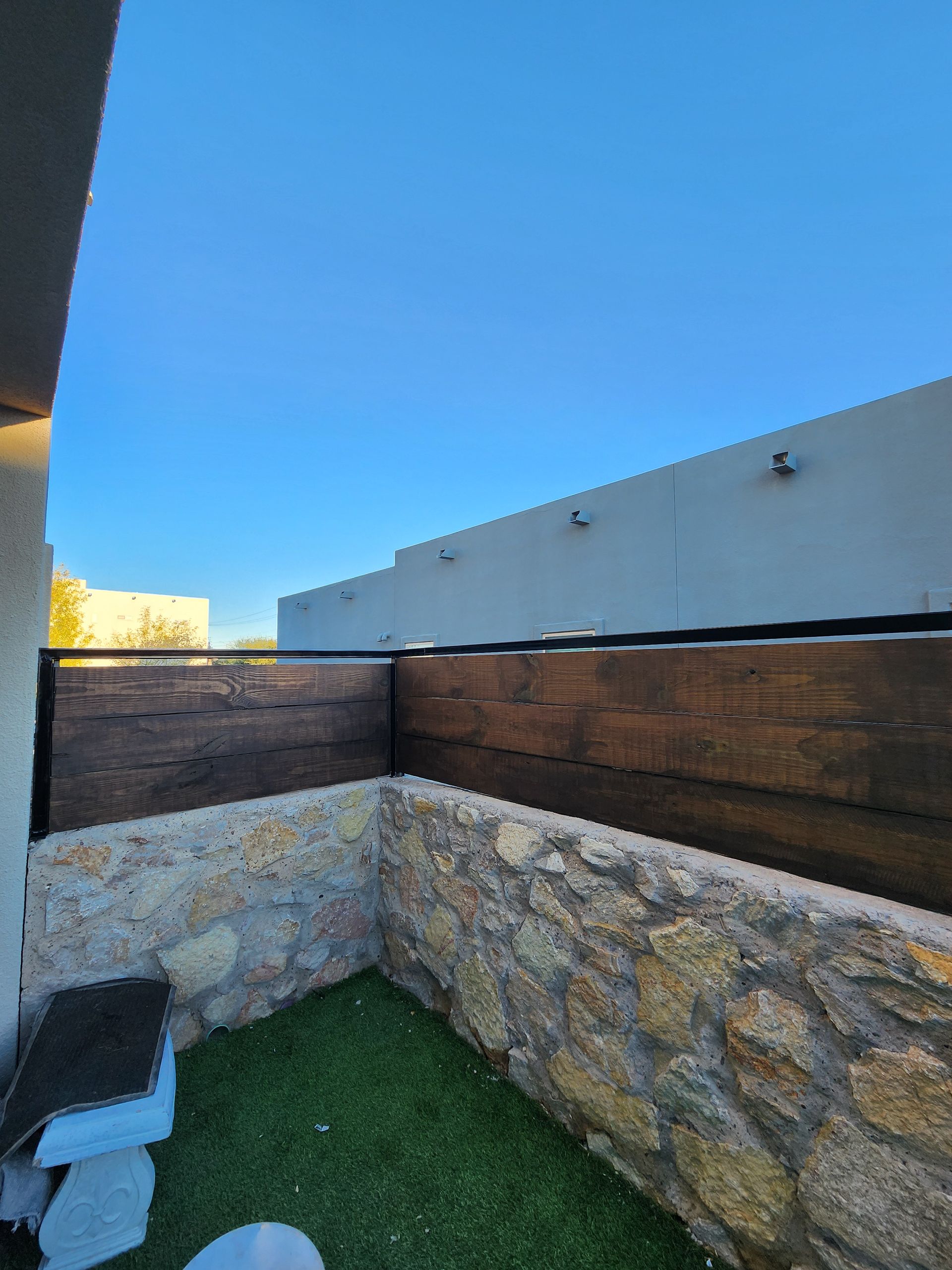 Small outdoor space with stone wall, grass, and wooden fence against a clear blue sky.