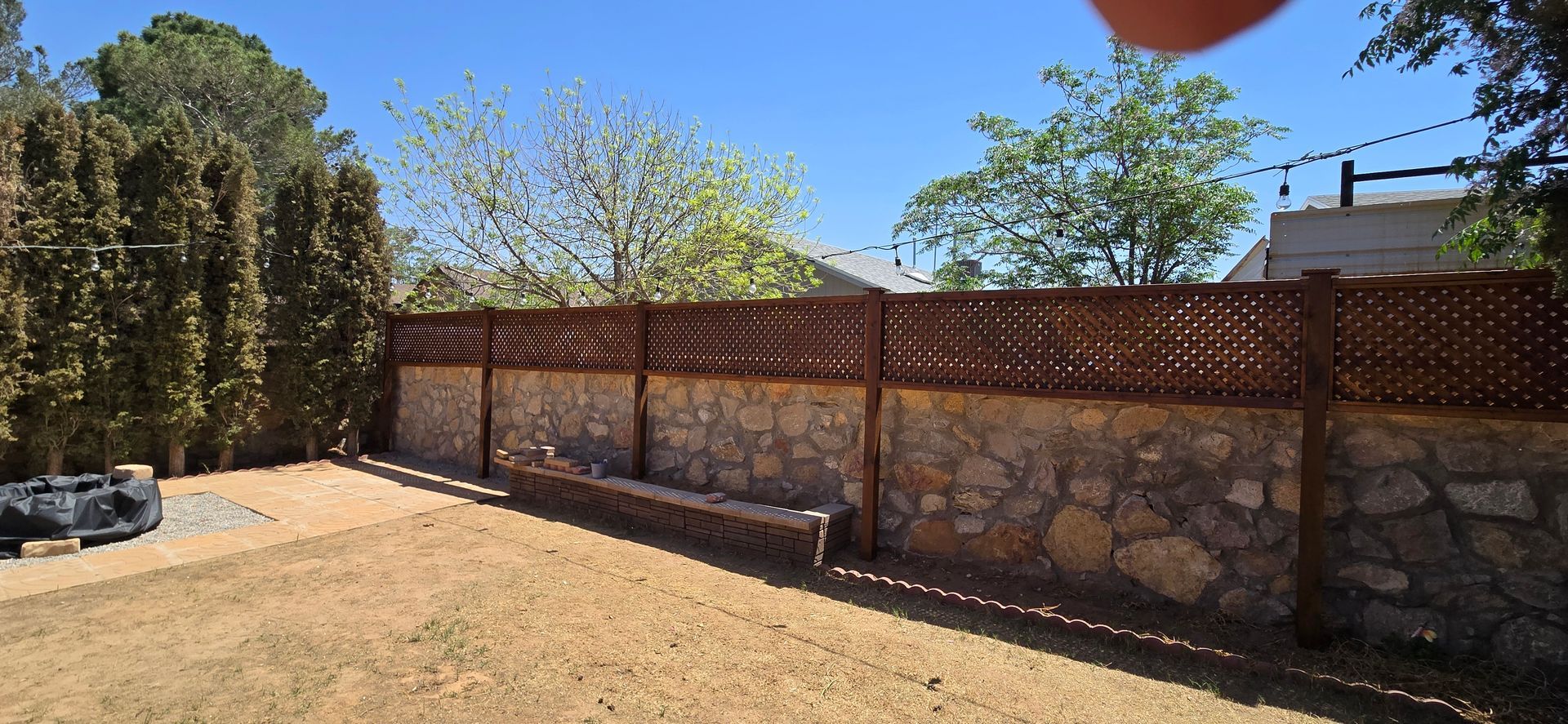 Backyard with a stone wall, wooden fence, and brown yard. Green trees and clear blue sky.