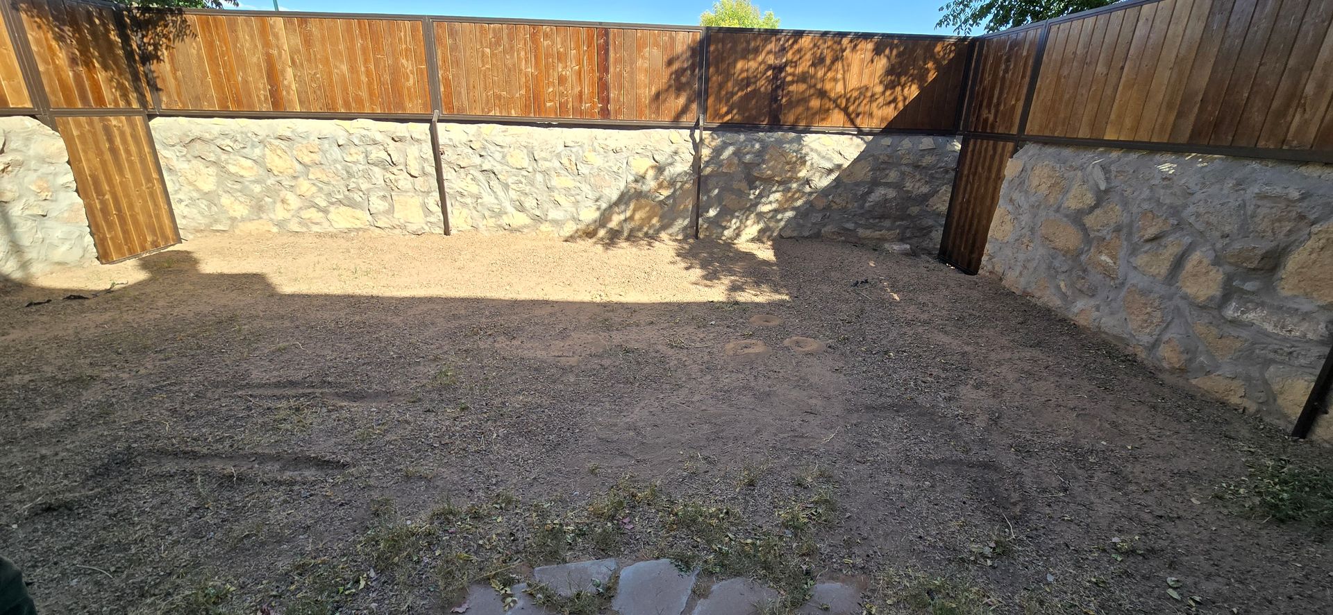 A backyard enclosed by a wooden fence and stone walls, with dry grass and gravel covering the ground.