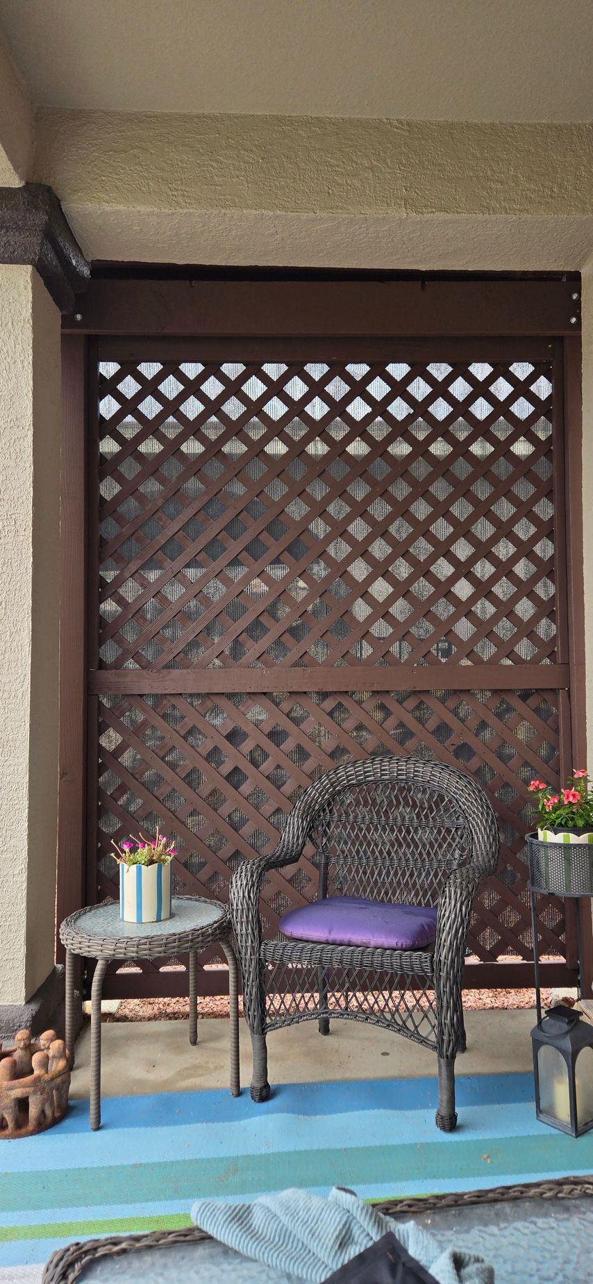 Wicker chair with purple cushion on a balcony, lattice wall behind. Table with flowers.
