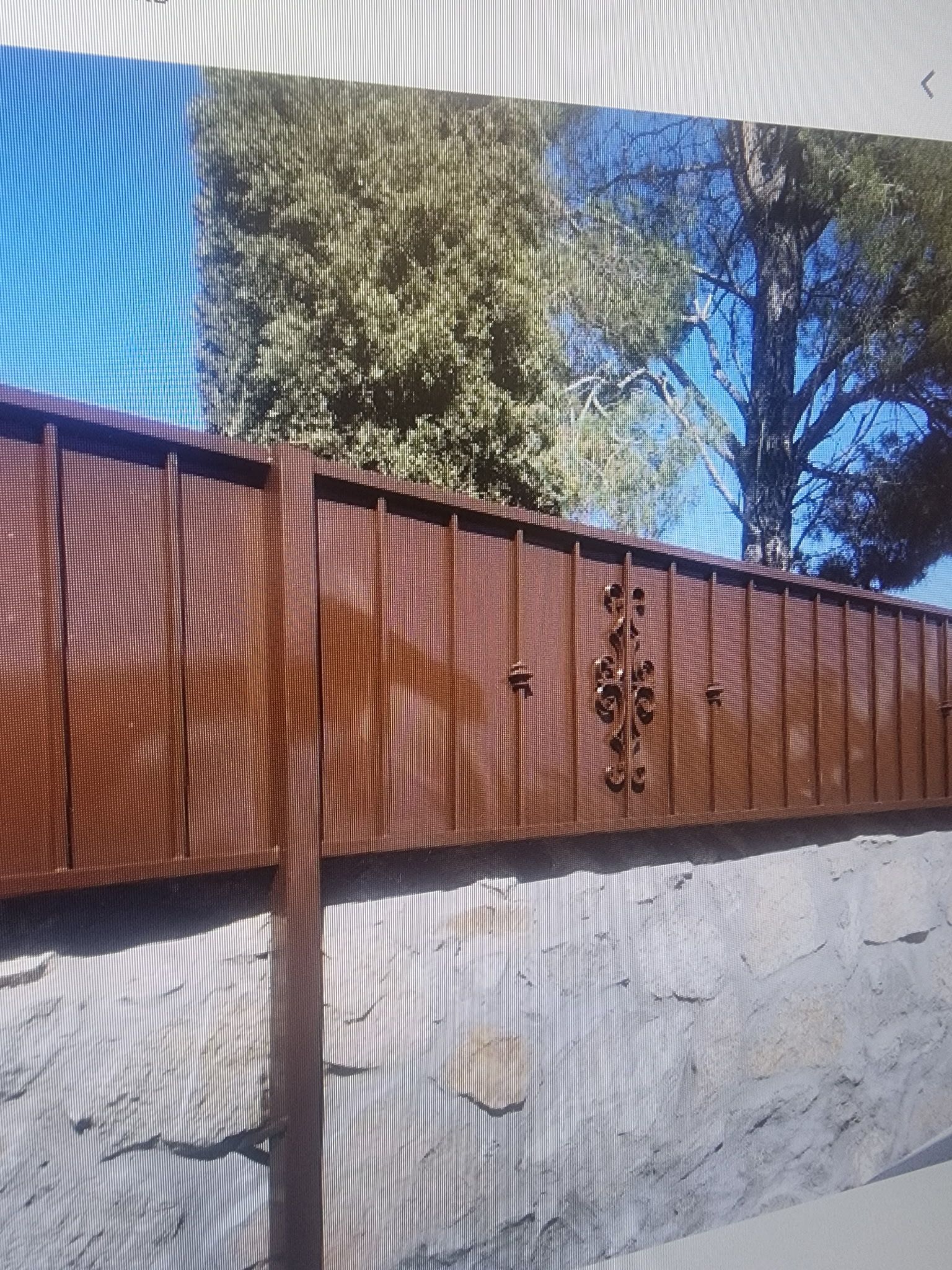 Brown metal fence with decorative detail on a gray stone wall, trees and blue sky in the background.