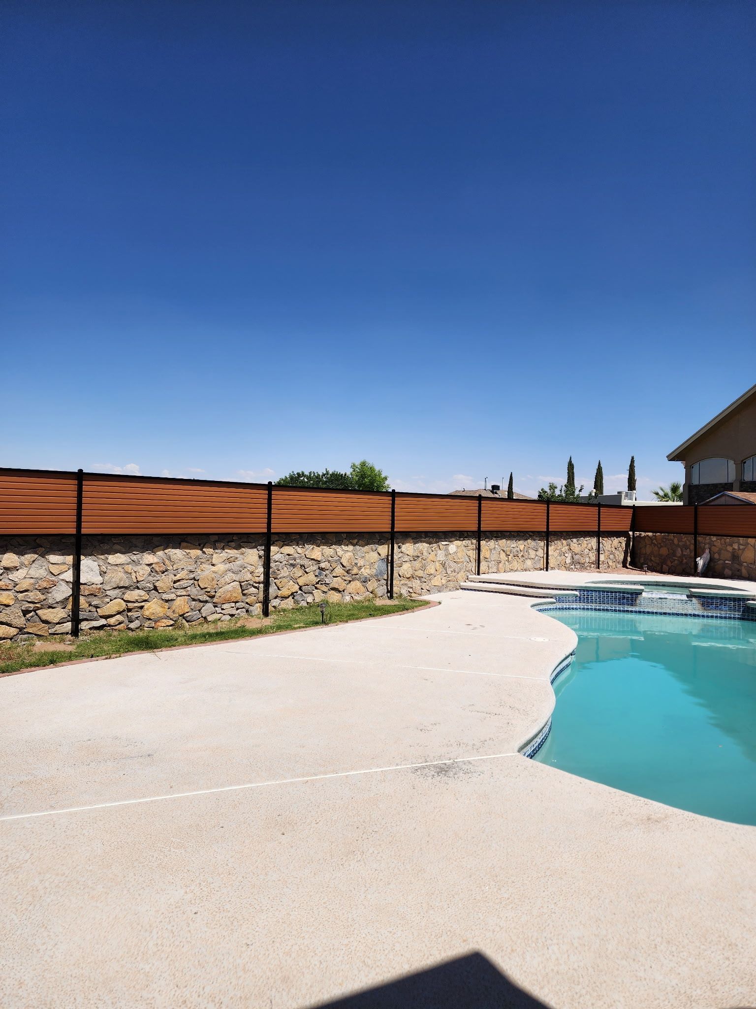 Poolside view with stone wall, black fence, and clear blue sky.