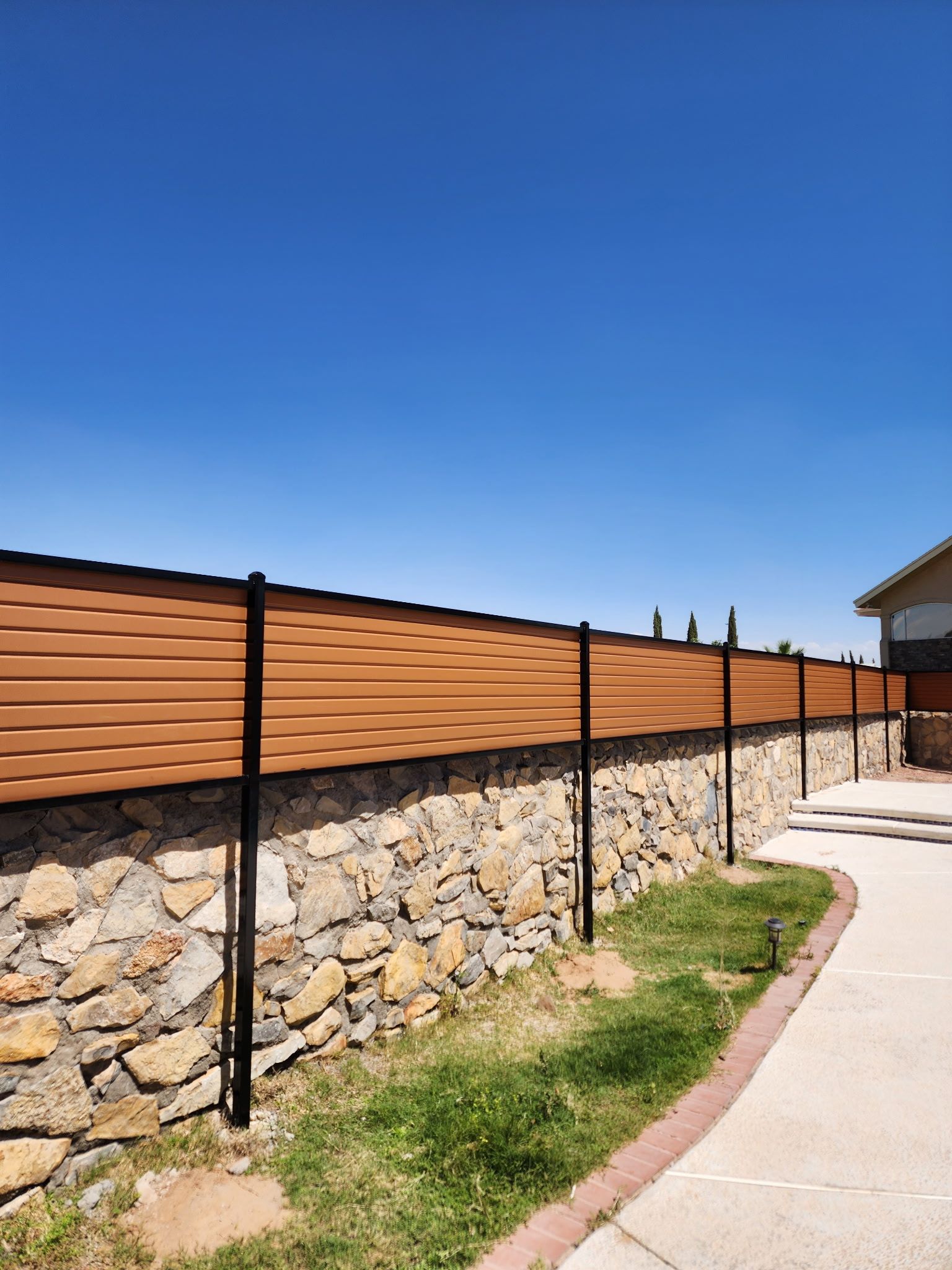 A brown and black horizontal fence sits atop a stone wall under a blue sky.