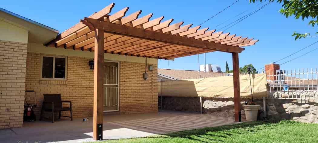 Wooden pergola attached to a light brick building on a sunny day. A chair sits nearby on the patio.