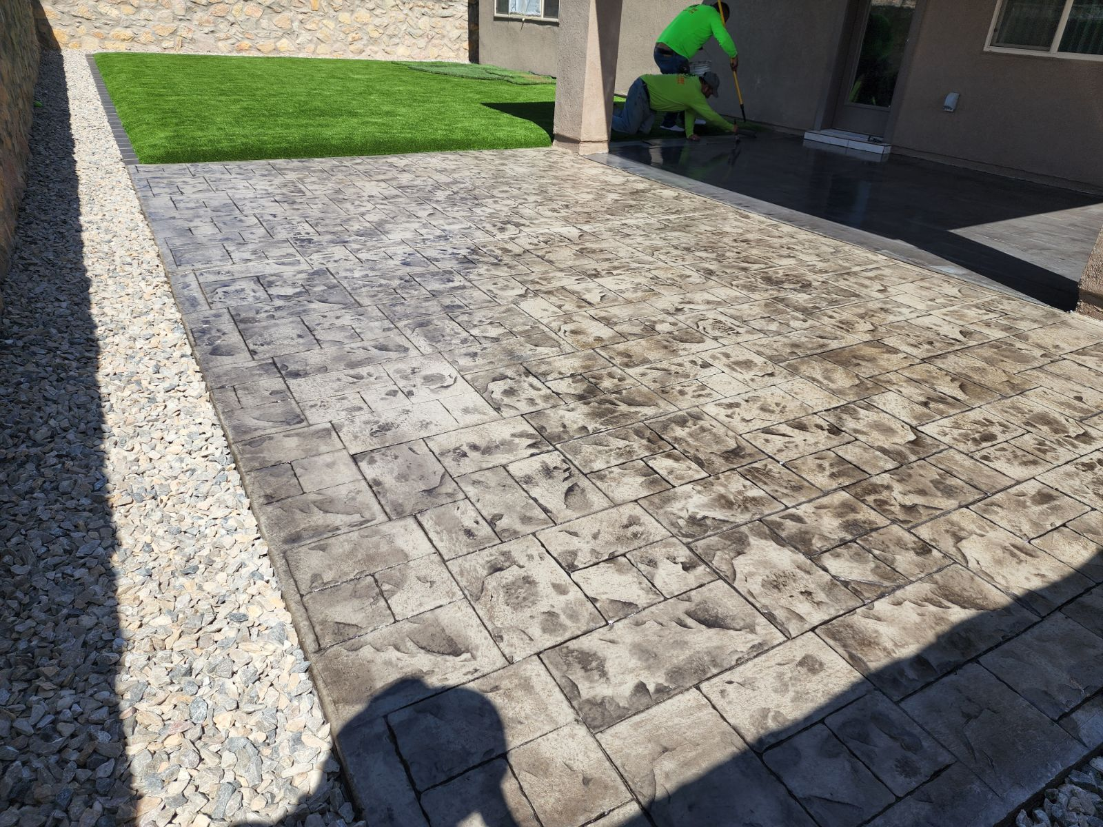 Workers installing gray stamped concrete patio with artificial turf section and gravel border.