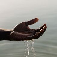 Water drips from a cupped hand held over a calm, light-colored body of water.