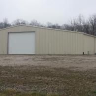 Tan industrial building with large white door, set on a gravel lot, with trees in the background.