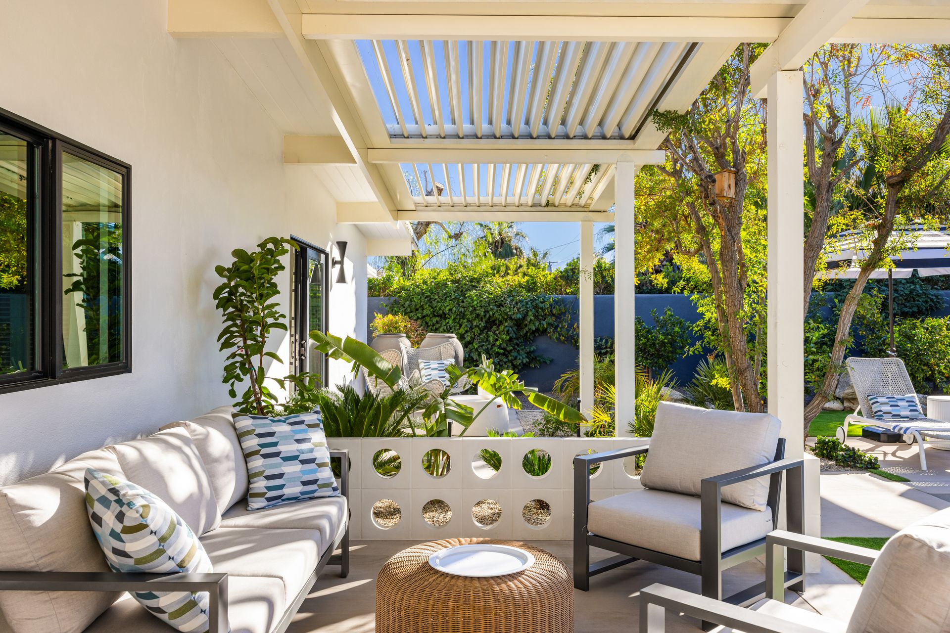 Outdoor patio with a couch, armchair, and woven ottoman. Bright, sunny setting with greenery.