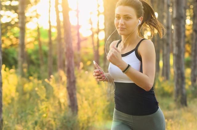 Woman jogging on a sunlit forest trail, holding a phone and wearing athletic clothes