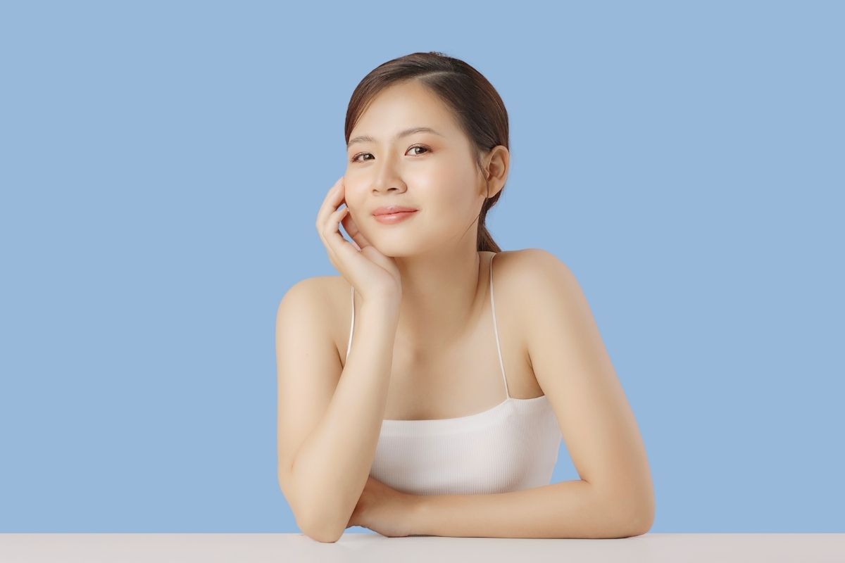Woman resting face on hand, smiling. Wearing white top, against light blue background.