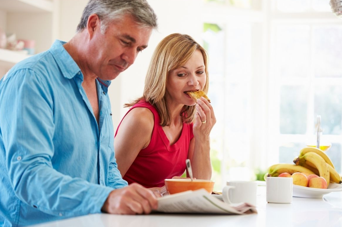 Two people eating breakfast at a kitchen table with fruit, coffee, and a newspaper