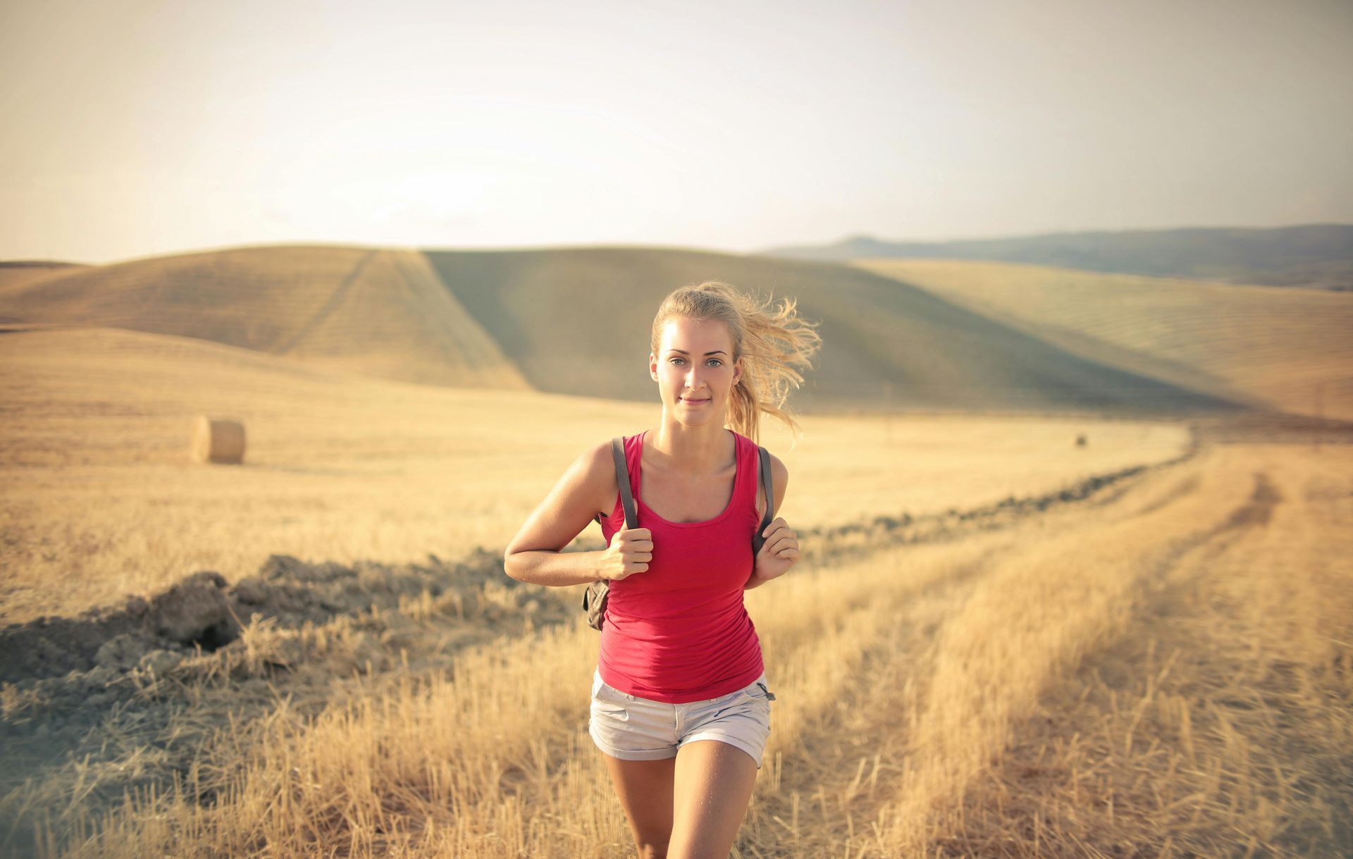 Blonde woman runs on a path through a field wearing red top and shorts, carrying a backpack, in a sunny landscape.