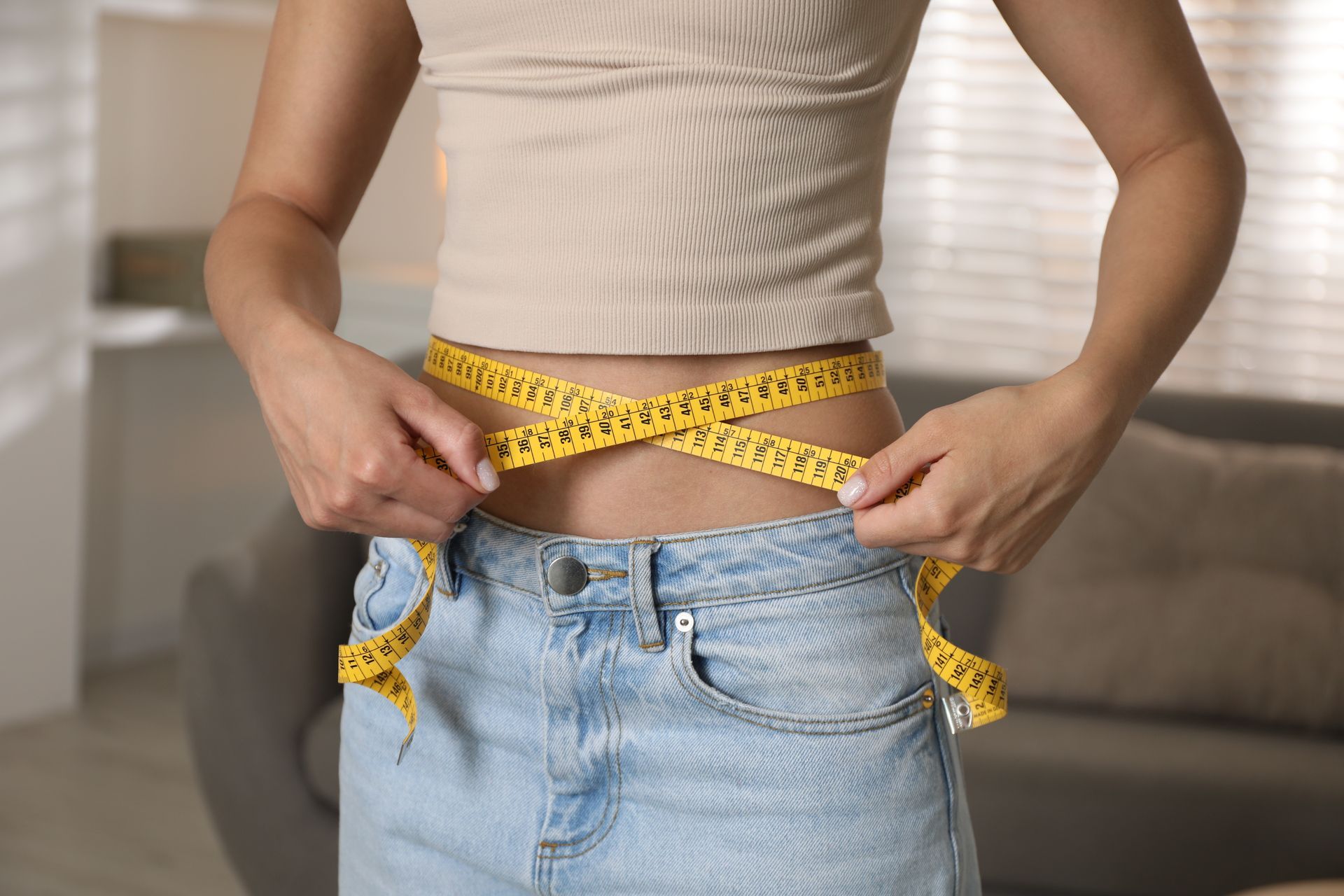 Woman measuring waist with yellow tape. In jeans and crop top.