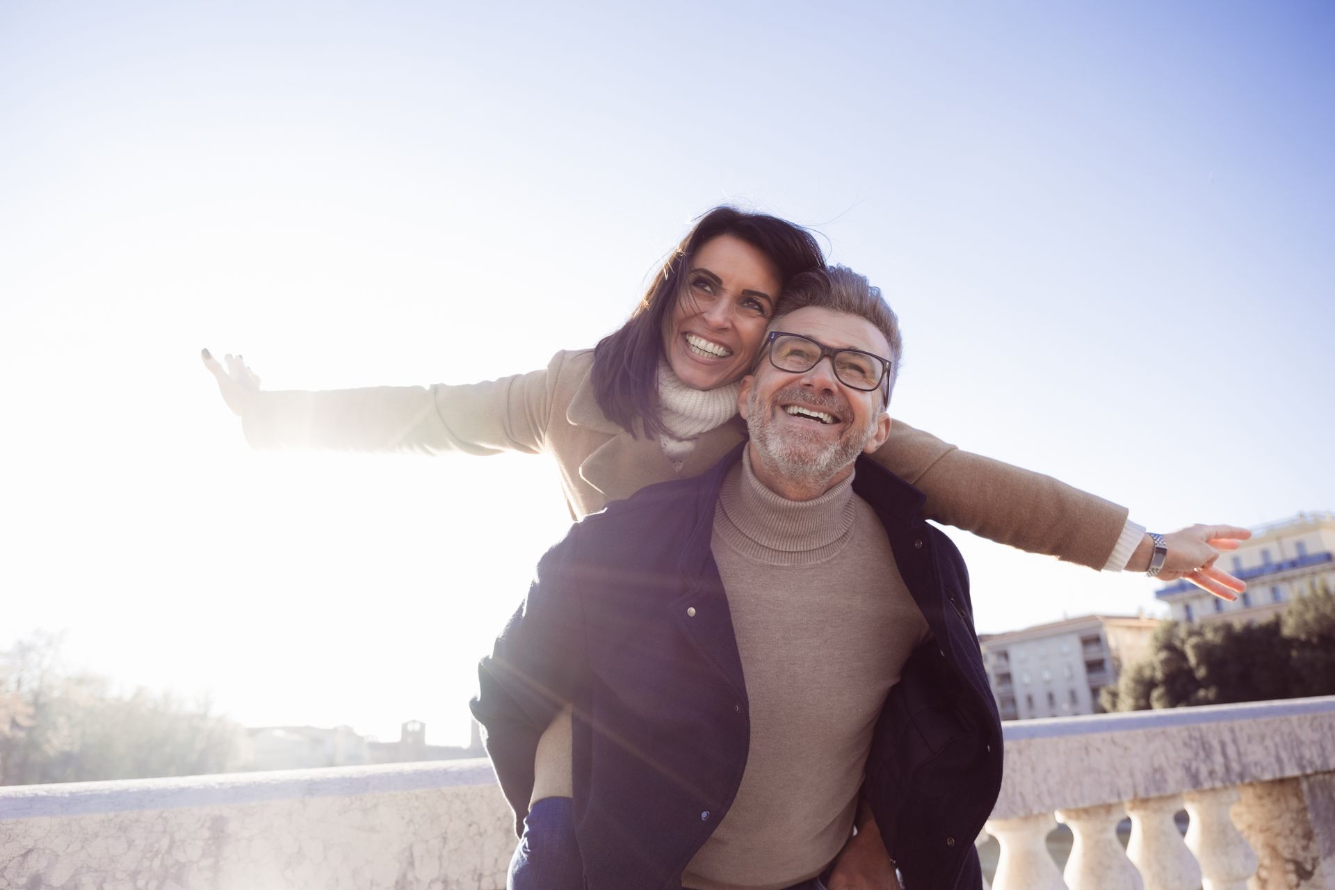Man giving woman a piggyback ride outdoors, both smiling with arms outstretched.