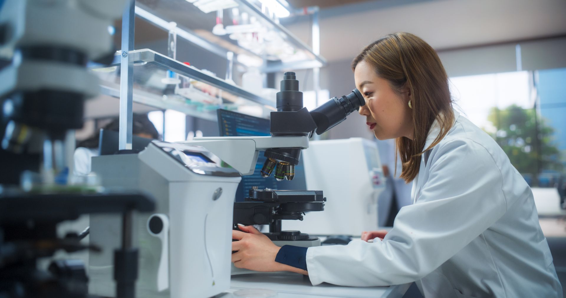 Woman in lab coat looks through a microscope in a laboratory setting.