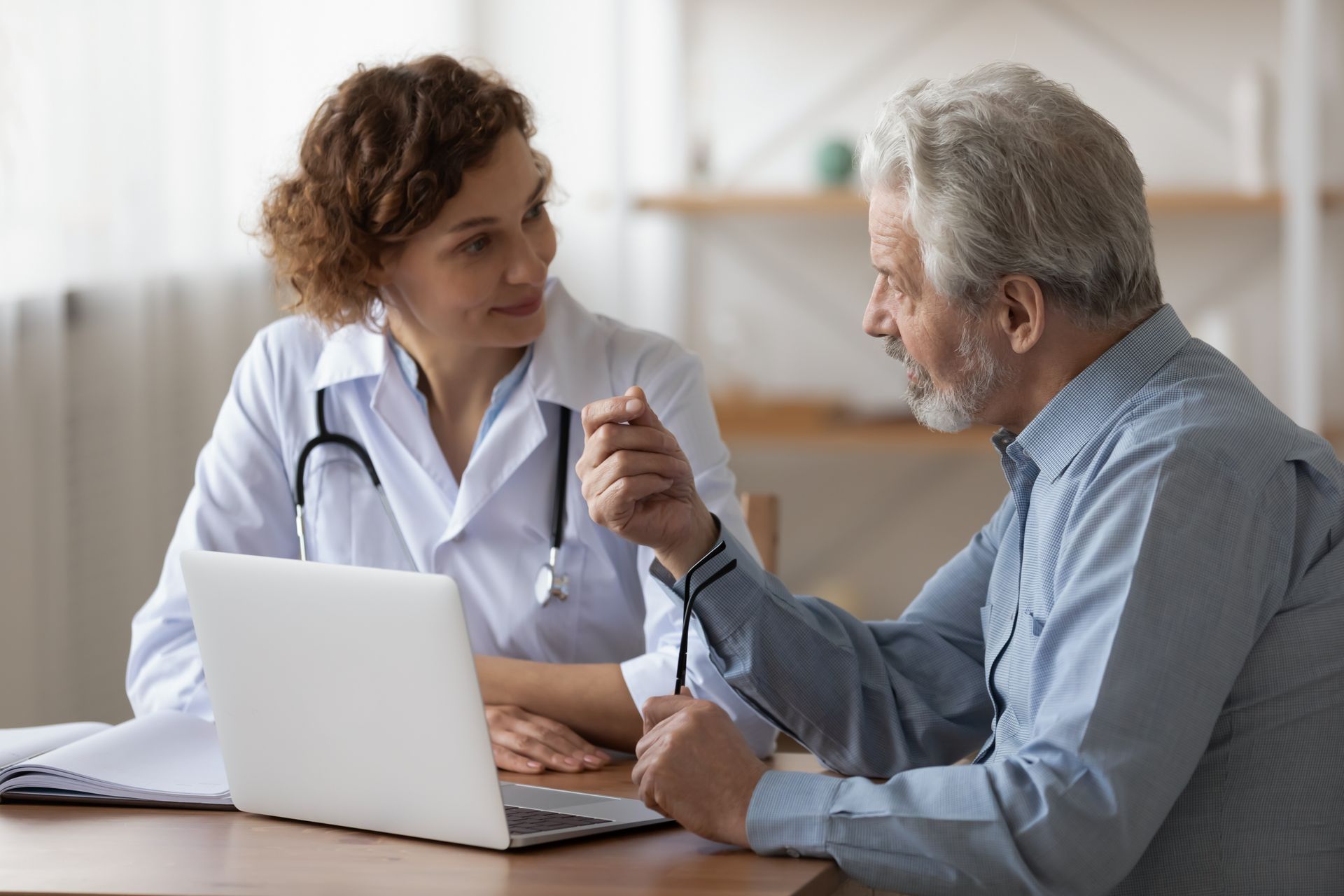 Doctor in white coat consults with a patient at a desk, looking at a laptop.