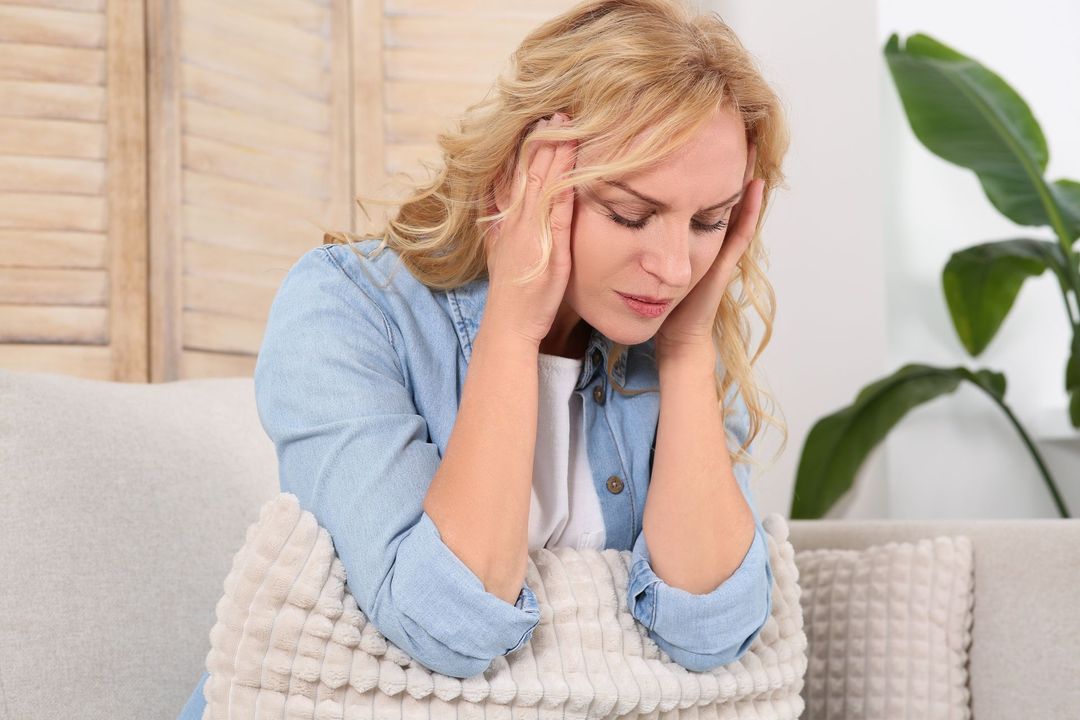 Woman with blonde hair holding her head, seated on a couch, looking distressed.
