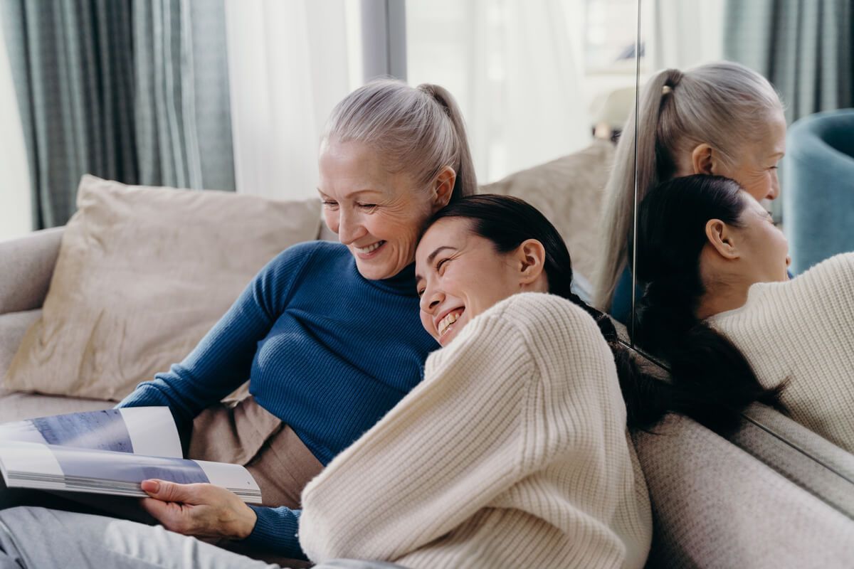 An elderly woman and a younger woman smile while looking at a book on a couch, leaning on a mirror.