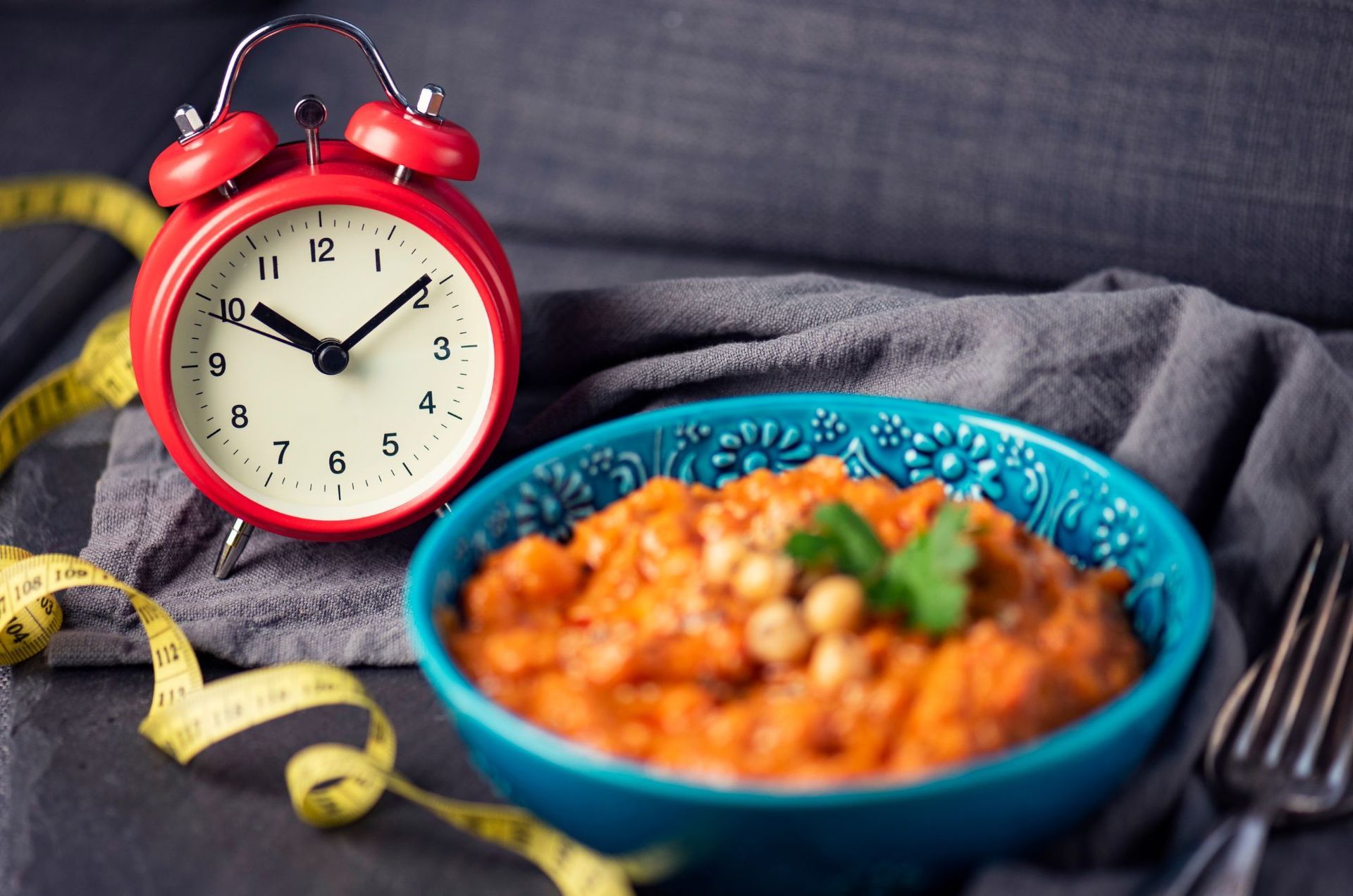 Red alarm clock, blue bowl of food, yellow measuring tape, and fork on a gray surface.