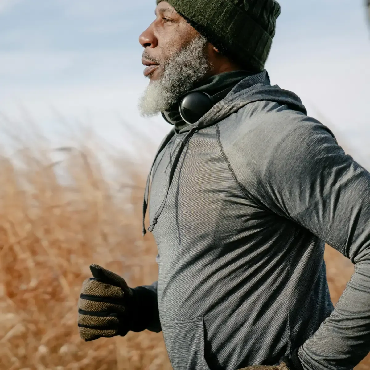 Man running outdoors, wearing grey hoodie, green hat, headphones, and gloves. Background of tall, brown grass.