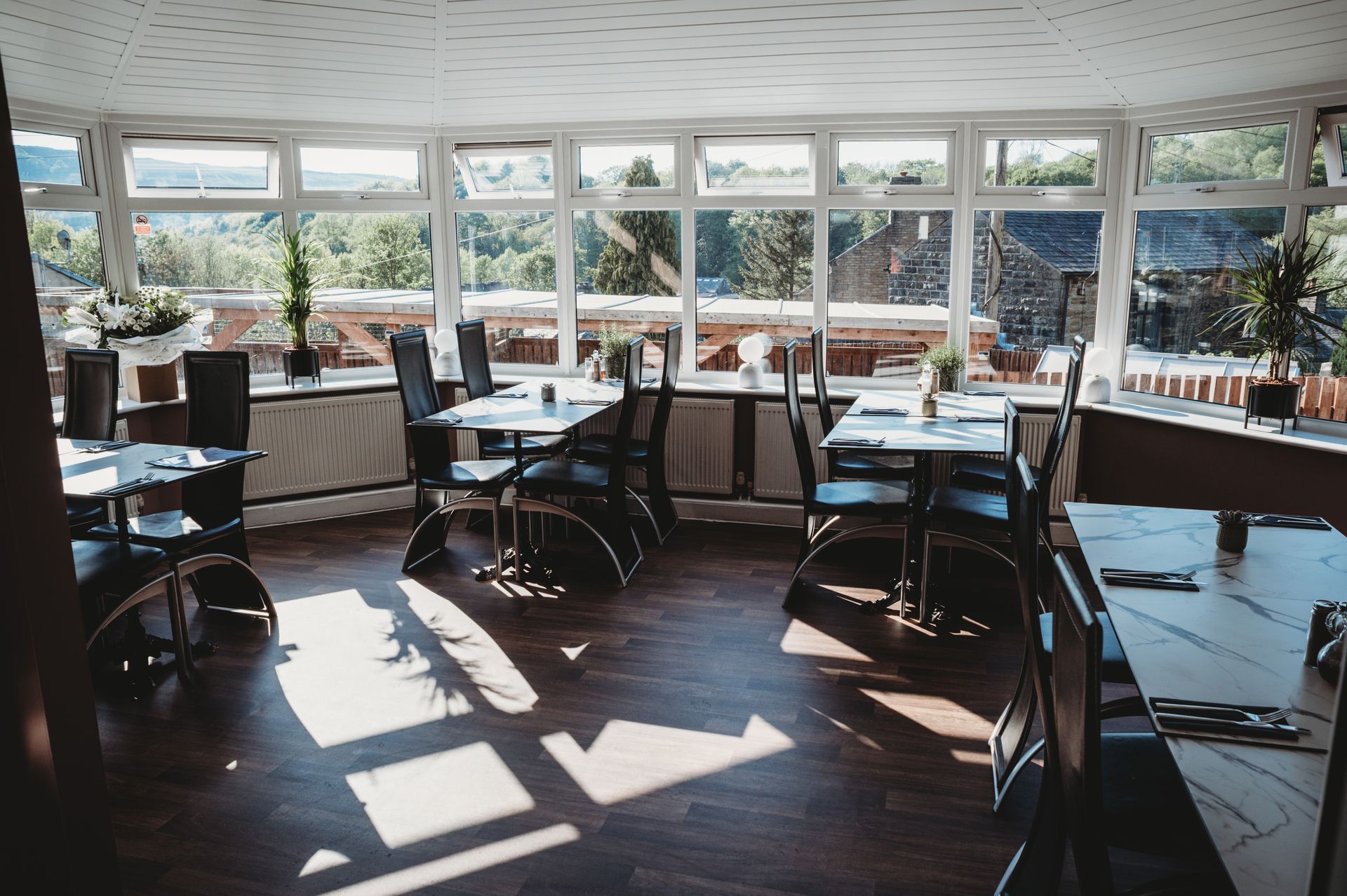 Restaurant dining room with tables and chairs near large windows, overlooking trees and a building.
