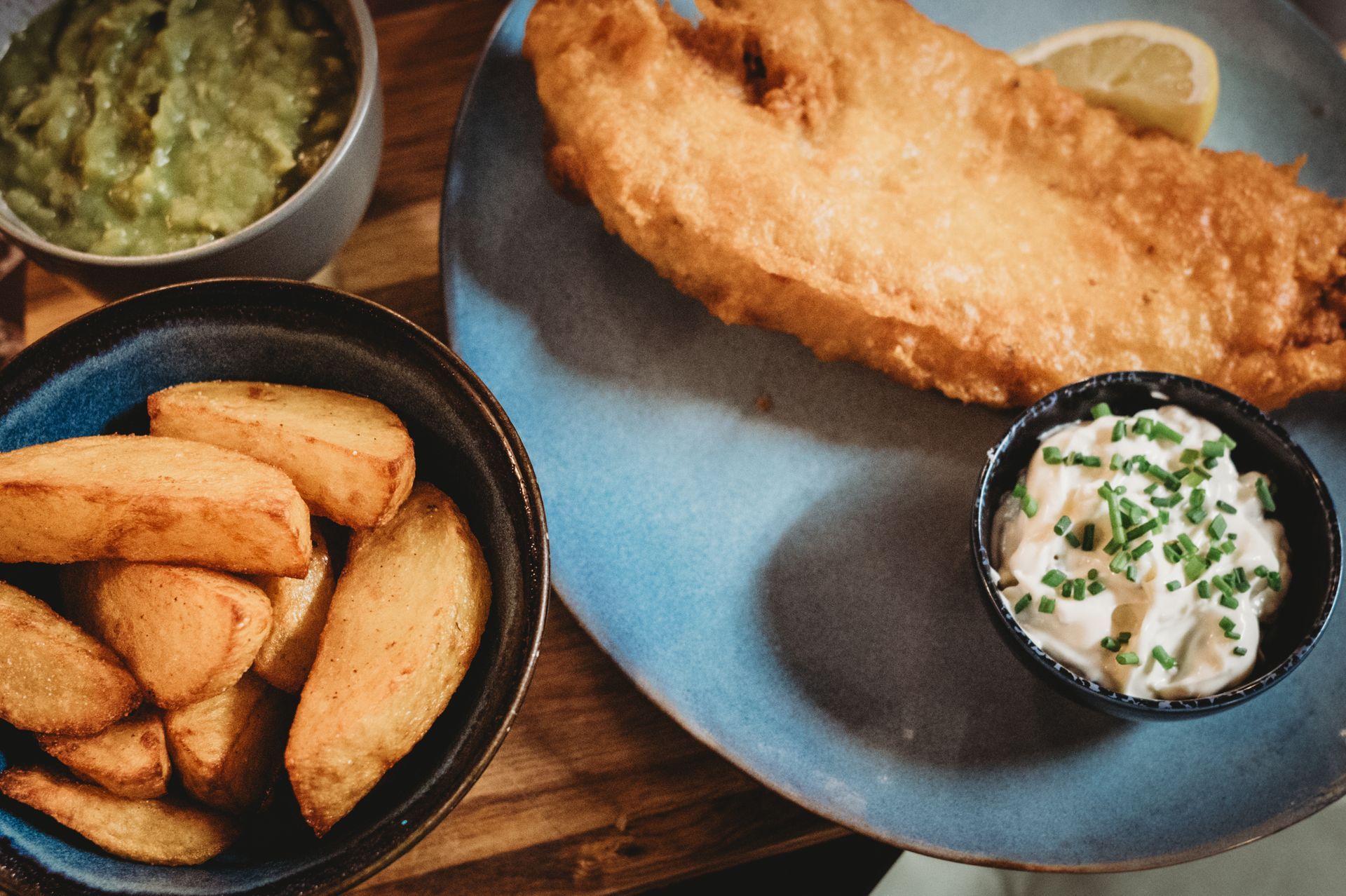Fish and chips with tartar sauce and guacamole on a blue plate, next to a bowl of potato wedges.