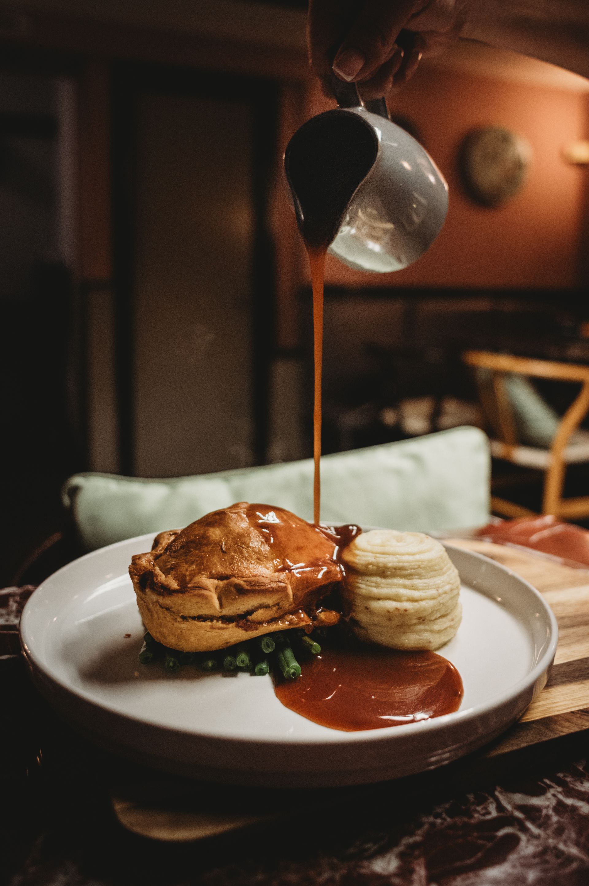A savory pie topped with gravy, next to a roll on a white plate. Sauce is being poured from above.