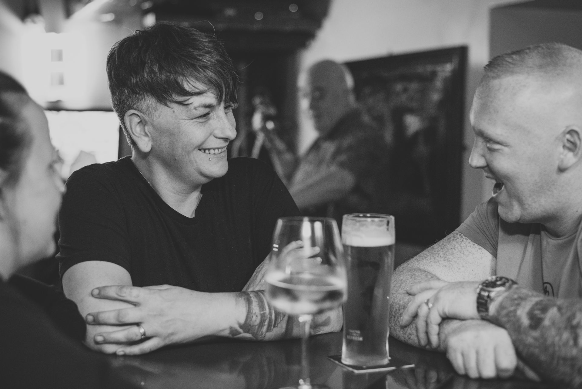 People smiling, at a bar. A woman with short dark hair in between two men with tattoos, glasses of drinks.