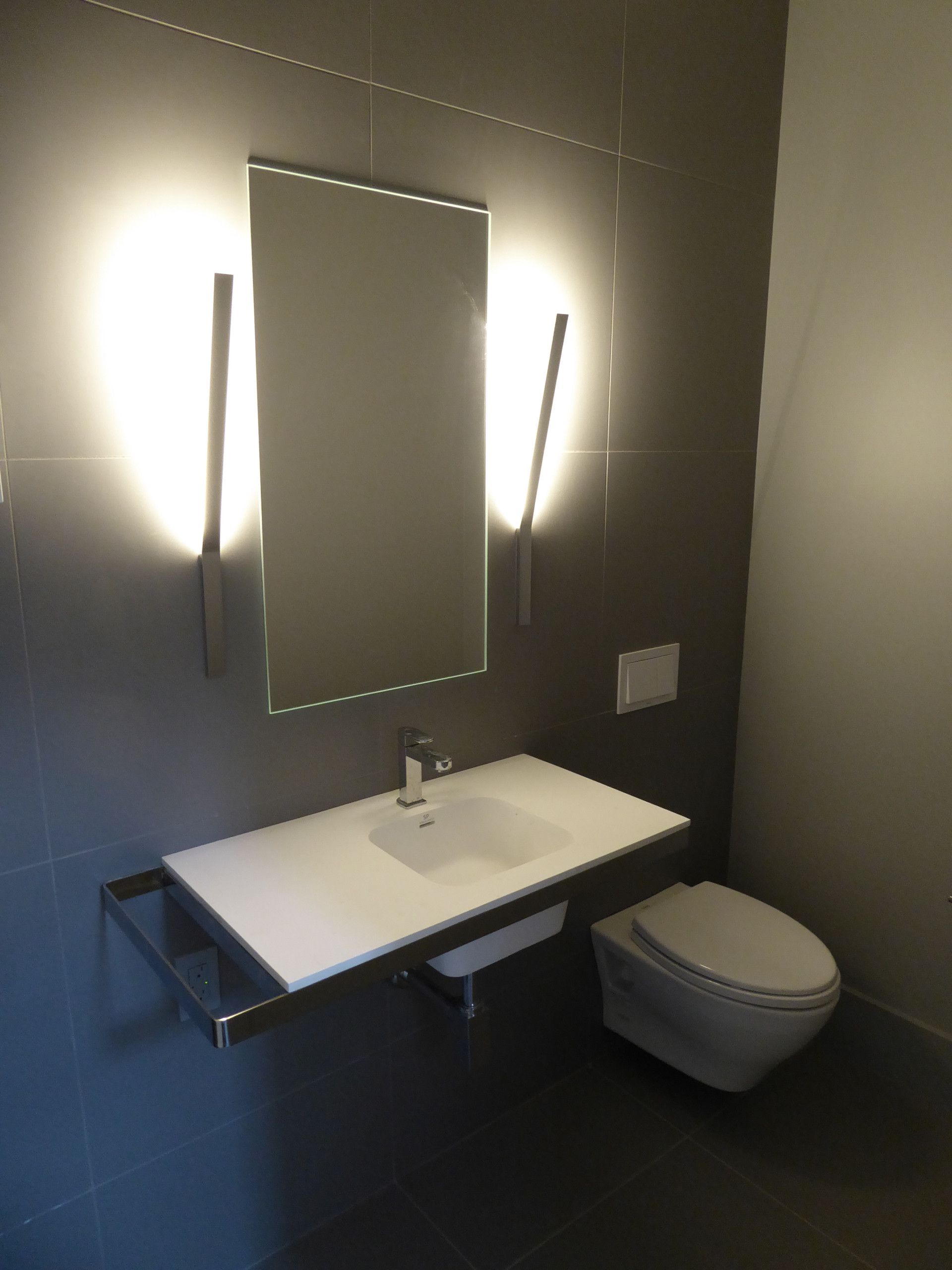 Modern bathroom with a floating sink, mirror, and toilet. Gray tiles and bright lighting.